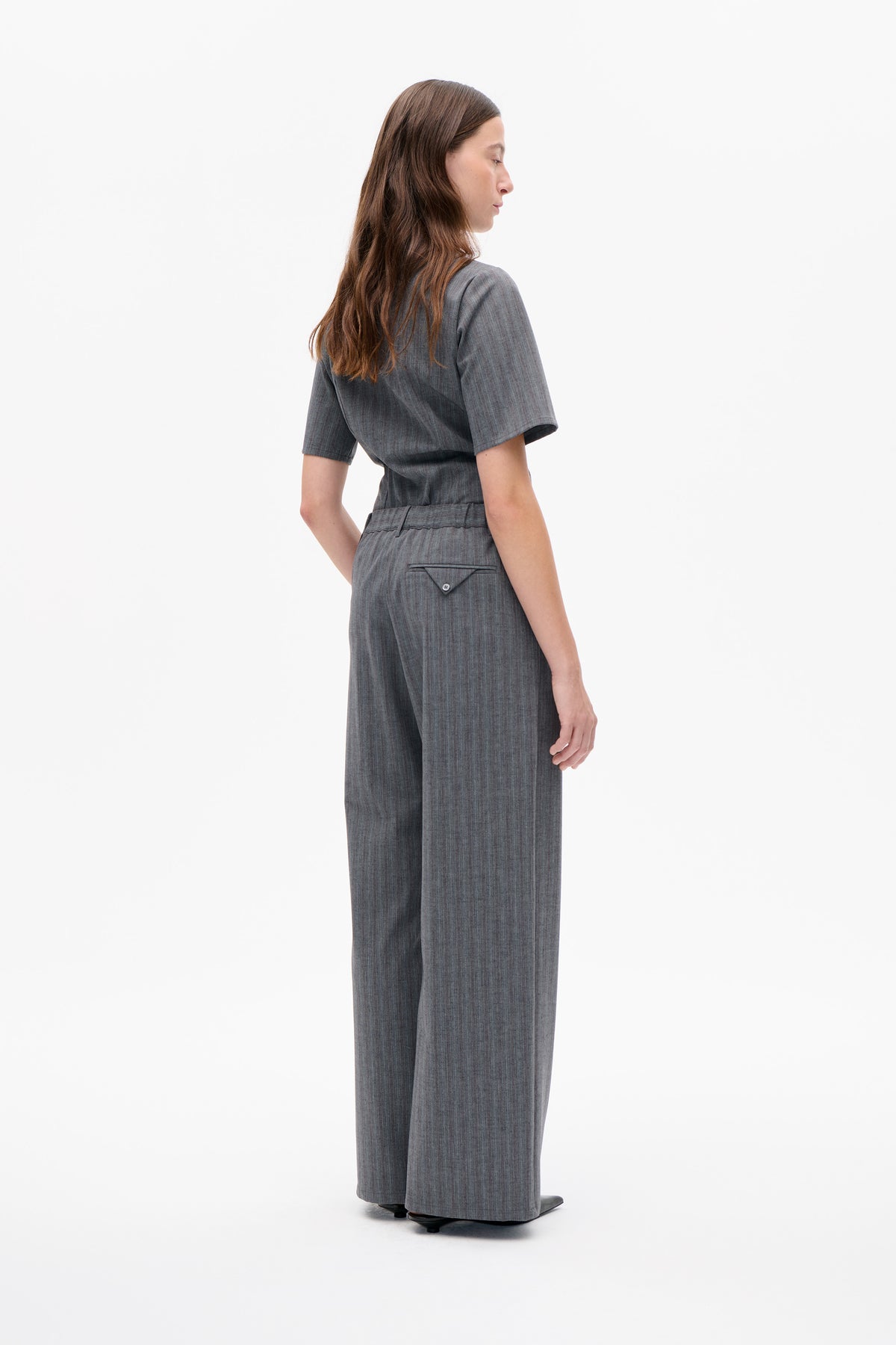 Side-back view of a charcoal grey pinstriped jumpsuit with short sleeves, wide legs and a buttoned back pocket.