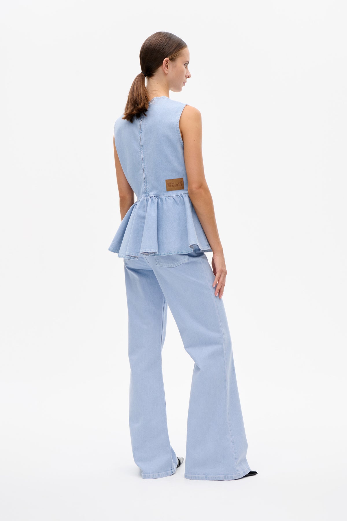 Back three-quarter view of a model wearing a light blue sleeveless denim peplum top with a logo patch and matching wide-leg jeans