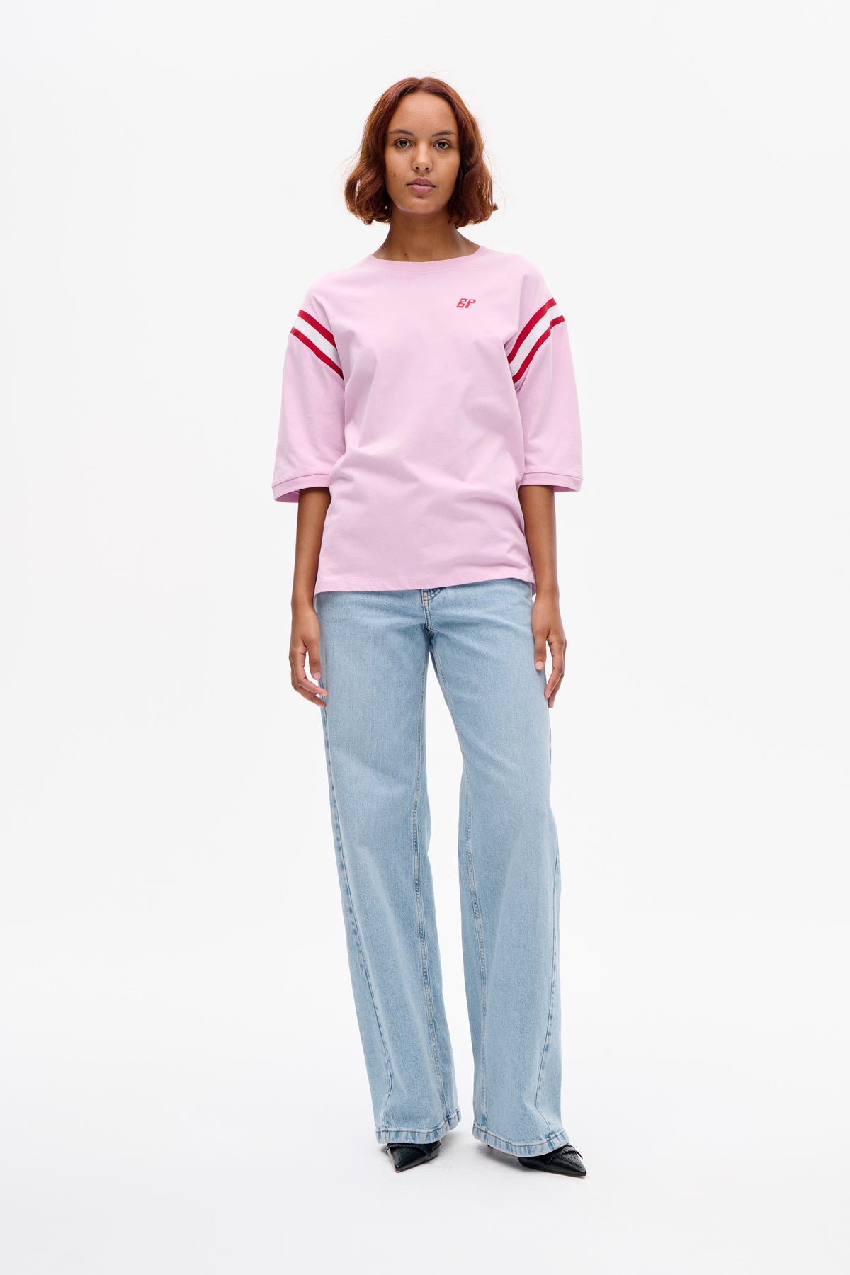Front view of a model wearing a light pink short-sleeved T-shirt with red sleeve stripes and a small chest logo, paired with light blue jeans.