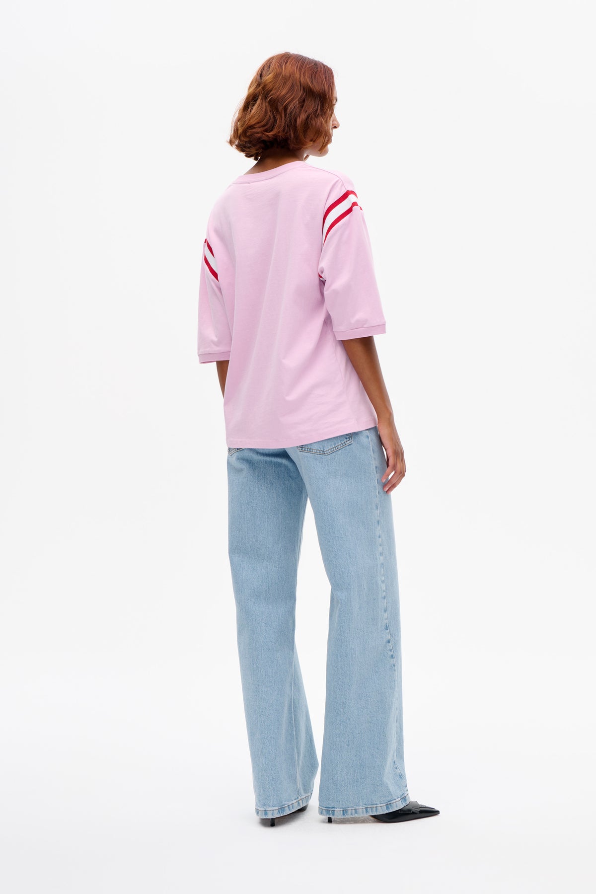 Back view of a model wearing a pale pink T-shirt with red and white sleeve stripes and light-wash wide-leg jeans.