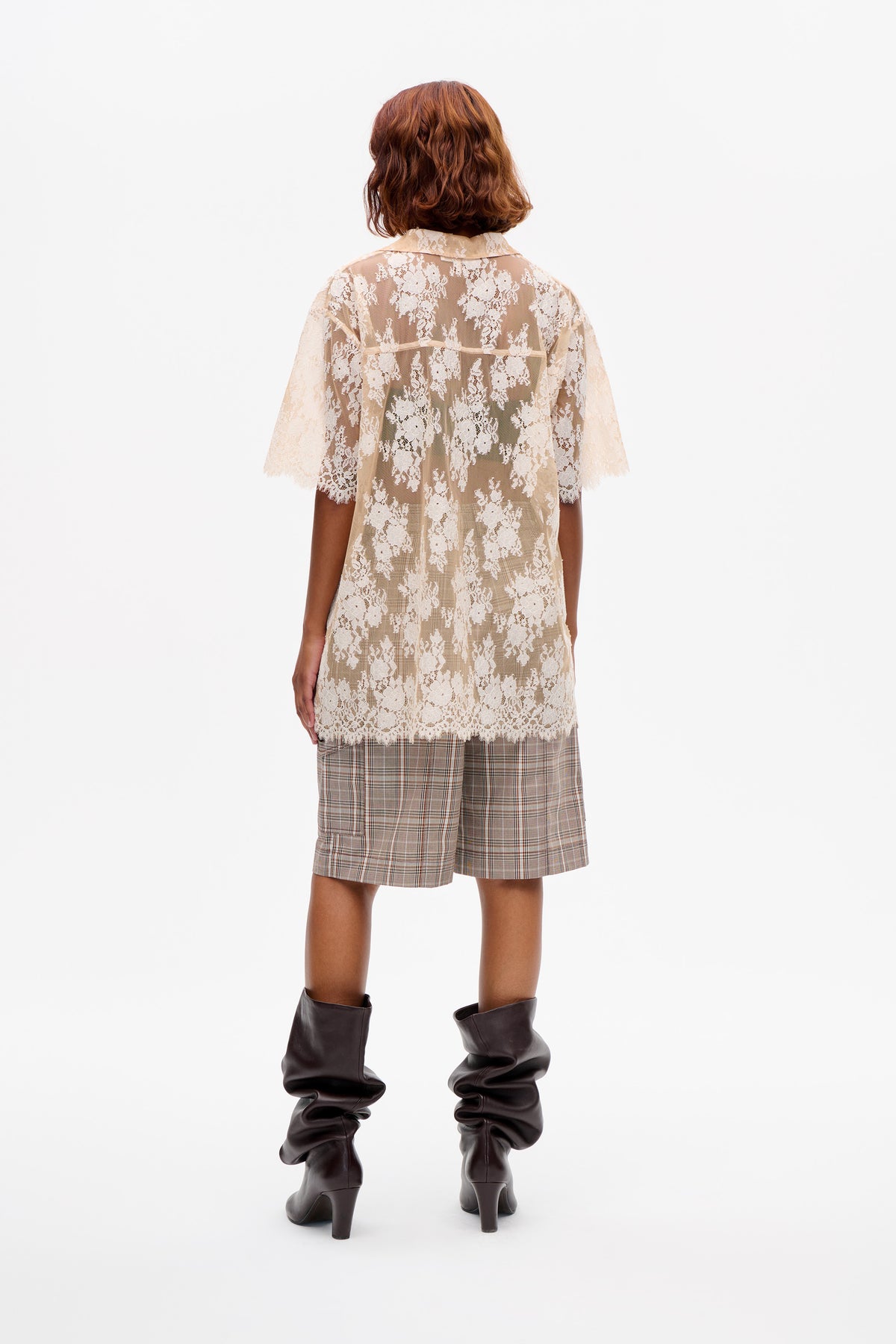 Back view of a model in a sheer cream floral lace collared tunic, brown plaid shorts and dark slouchy heeled boots.
