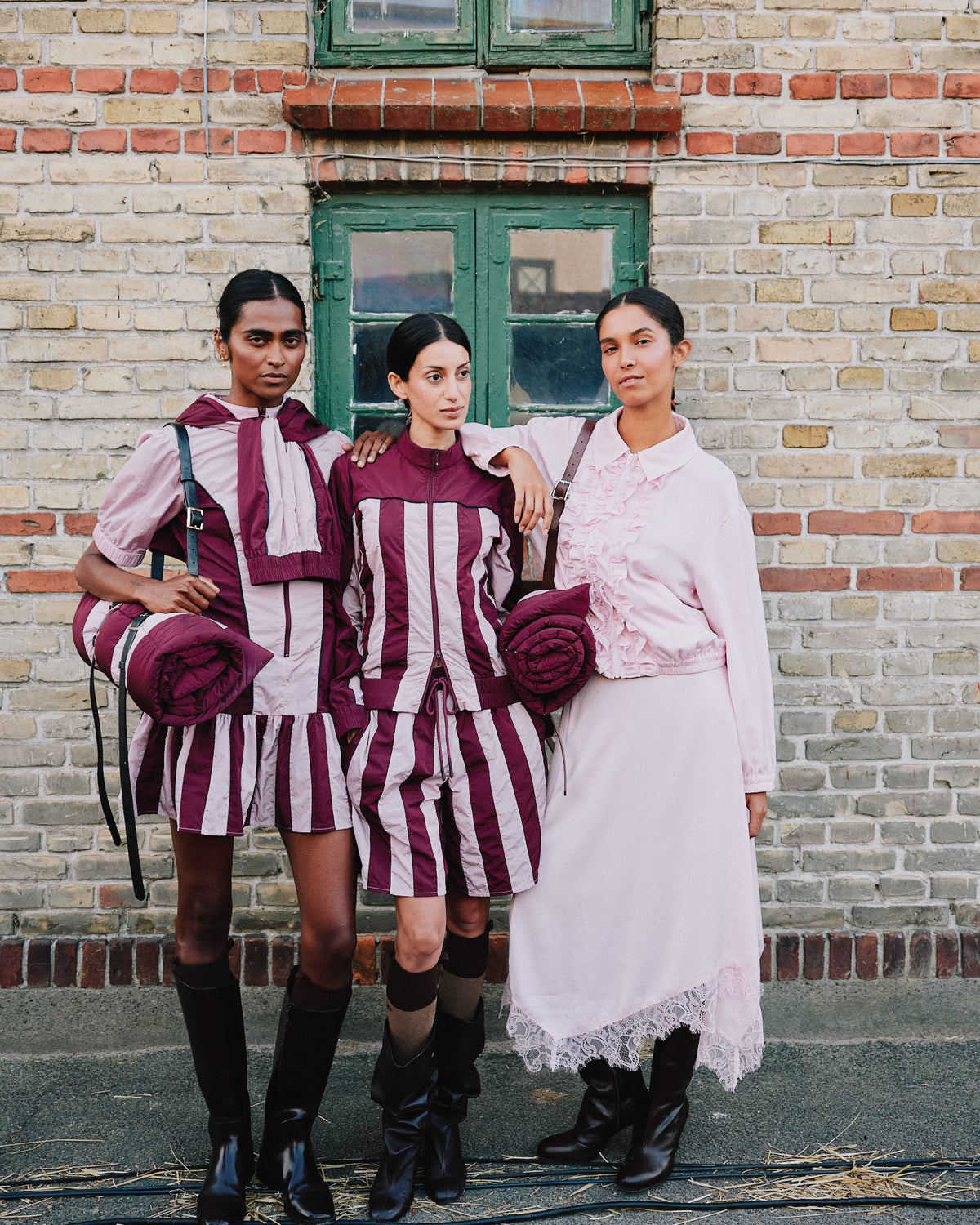 Three models pose outside in plum-and-pale-pink striped outfits and a pale pink ruffled dress, carrying rolled matching garments.