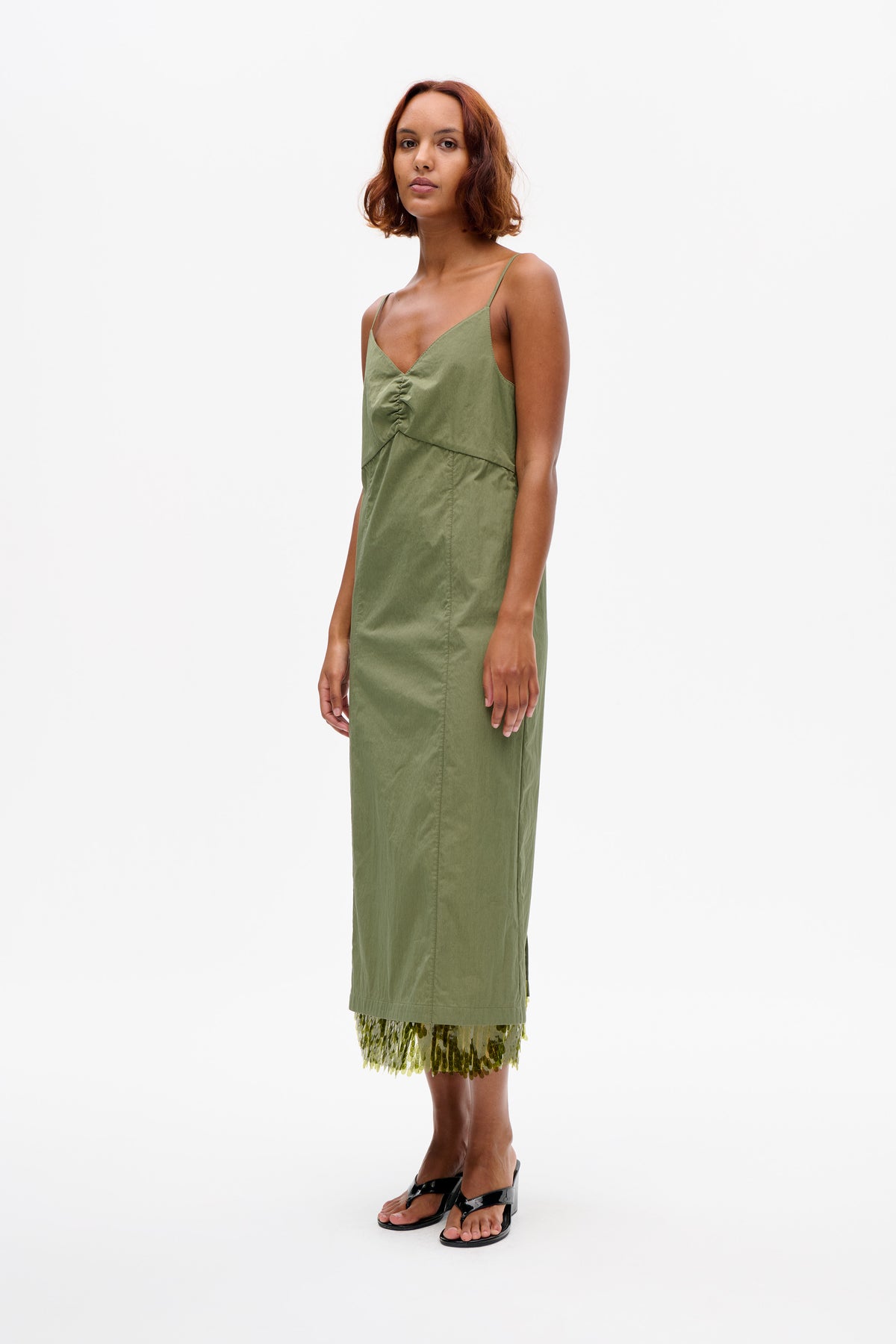 Model wearing a sage green spaghetti-strap dress with a V-neck and gathered front, layered over a green sequin skirt.