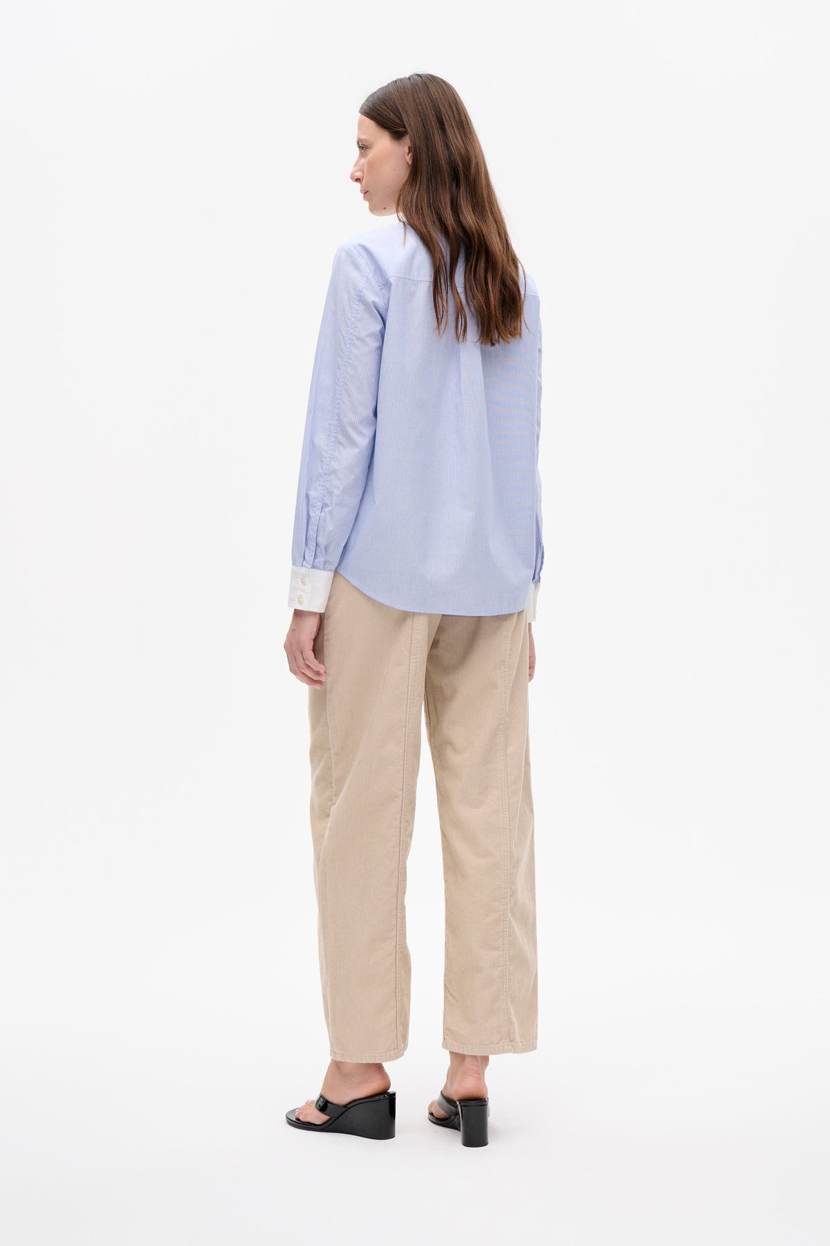 Model shown from a rear three-quarter view wearing a pale blue shirt with fine vertical stripes and white cuffs, paired with beige trousers.