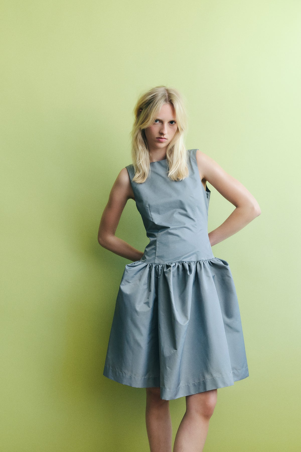 Model poses against a pale green backdrop wearing a sleeveless blue-grey dress with a gathered waist and full skirt.