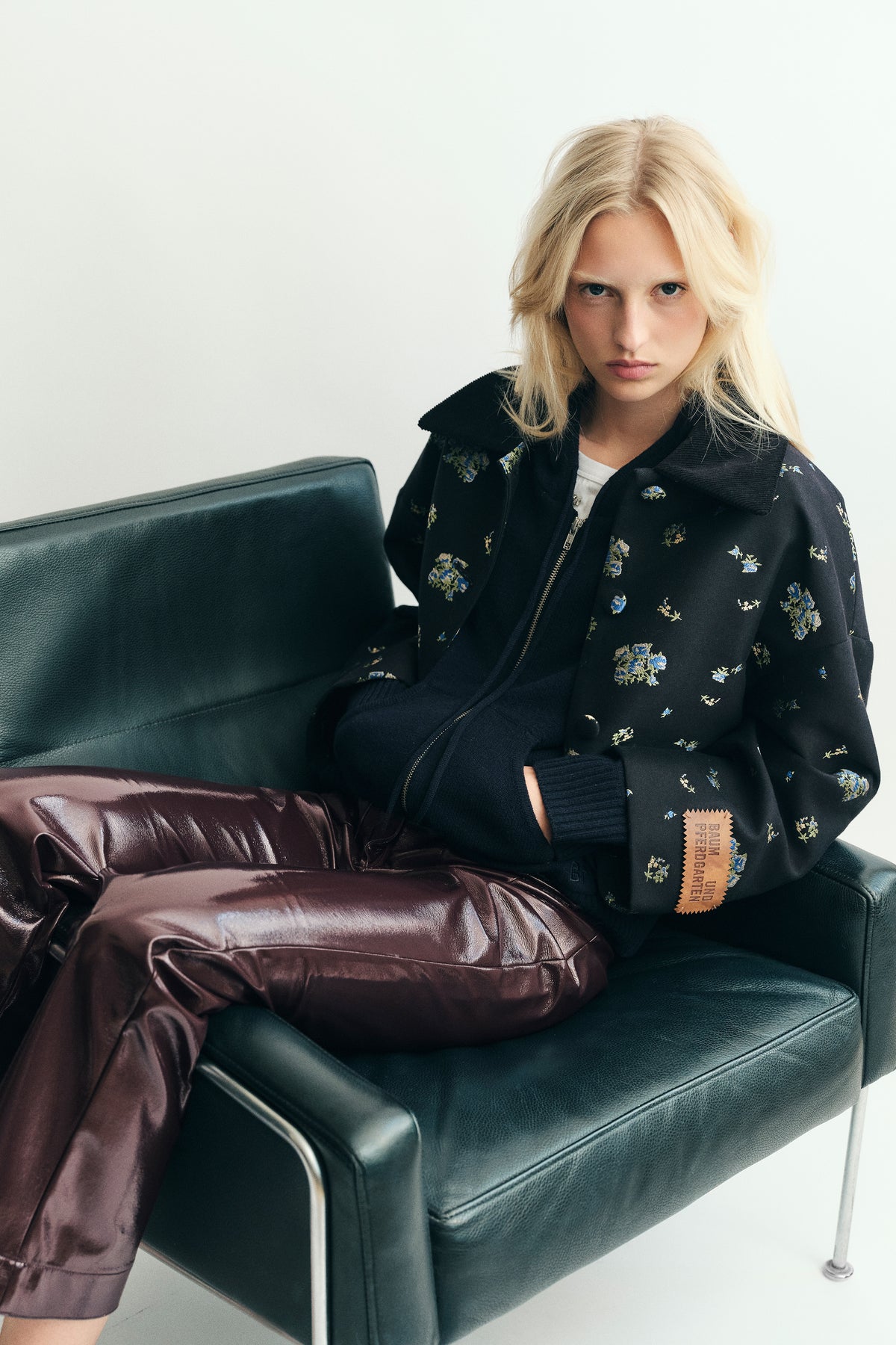 Seated model wearing a black zip-front jacket with small blue and yellow floral motifs and glossy burgundy trousers.