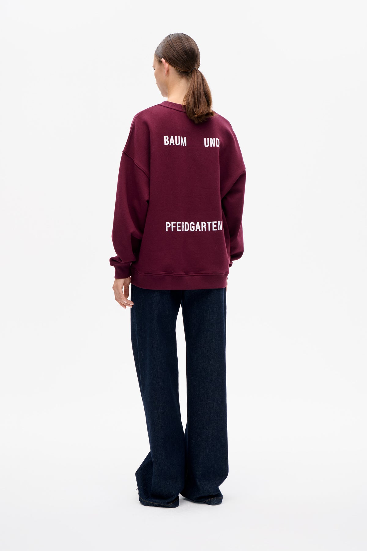 Back view of a model wearing a burgundy oversized sweatshirt with white Baum und Pferdgarten lettering and dark wide-leg jeans.