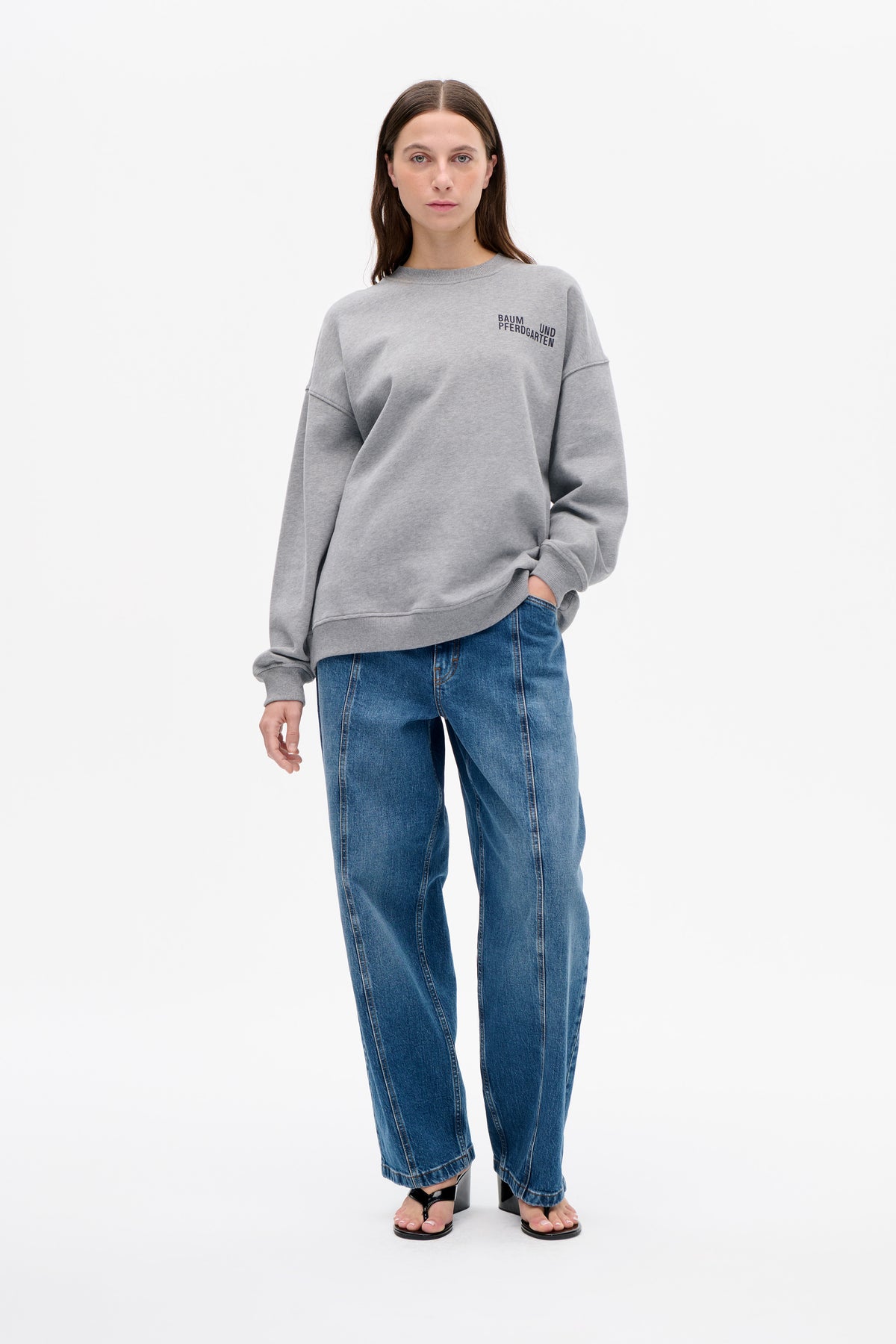 Front view of a model wearing a loose grey sweatshirt with ribbed trims and a small Baum und Pferdgarten chest logo, paired with blue jeans.