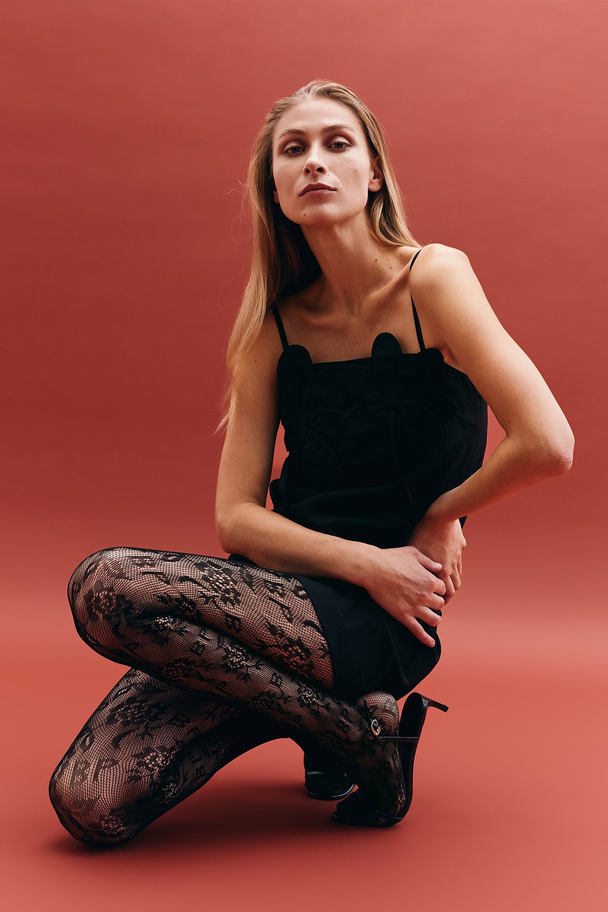 A seated model wears a black sleeveless dress with a sculptural floral bodice, lace-patterned tights and black heels against a coral backdrop.