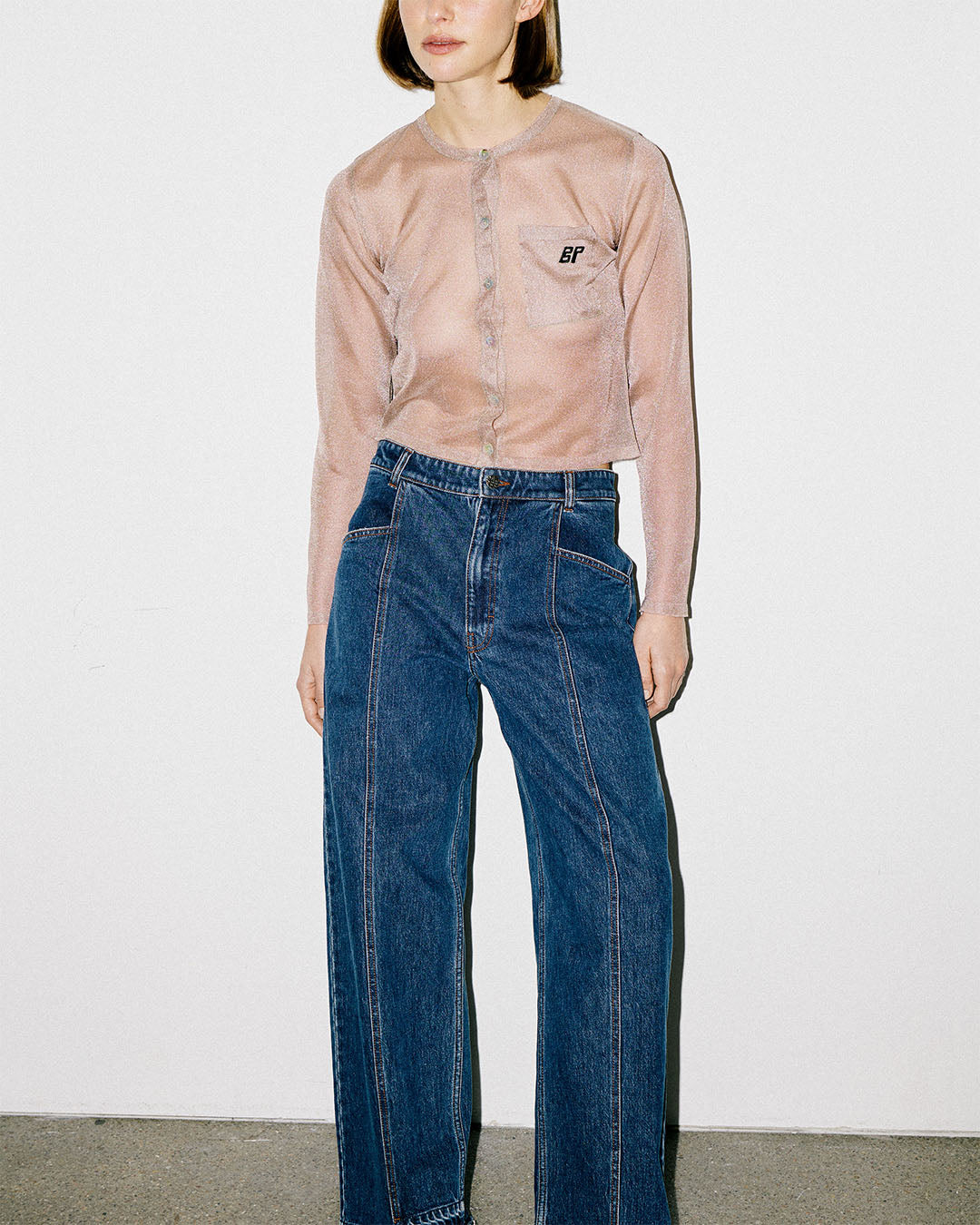 Front view of a model wearing a sheer blush-beige button-front shirt with a chest pocket and dark blue wide-leg jeans.