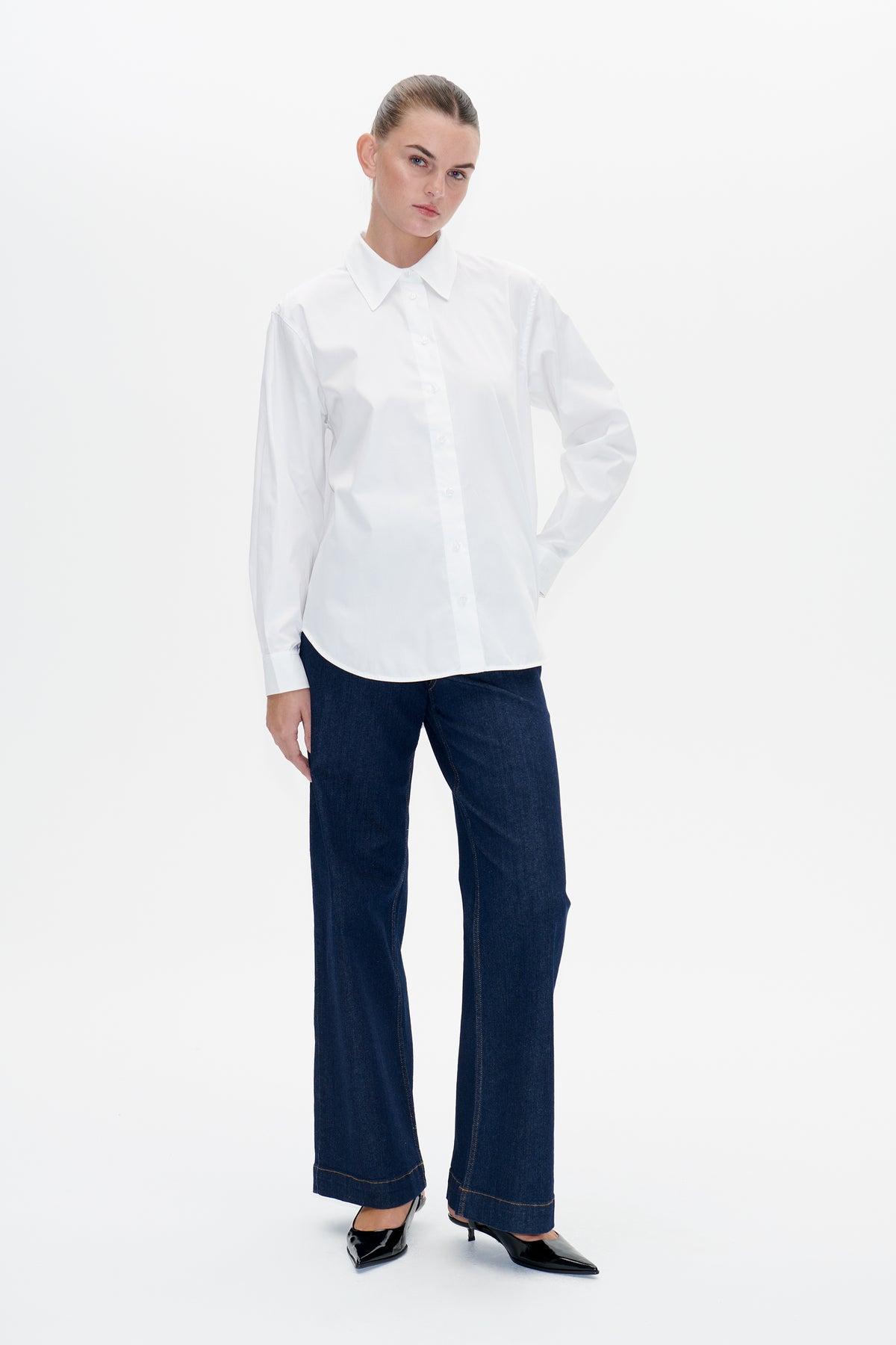 Model wearing a white long-sleeved button-up shirt with a pointed collar and dark blue wide-leg jeans, shown from the front.