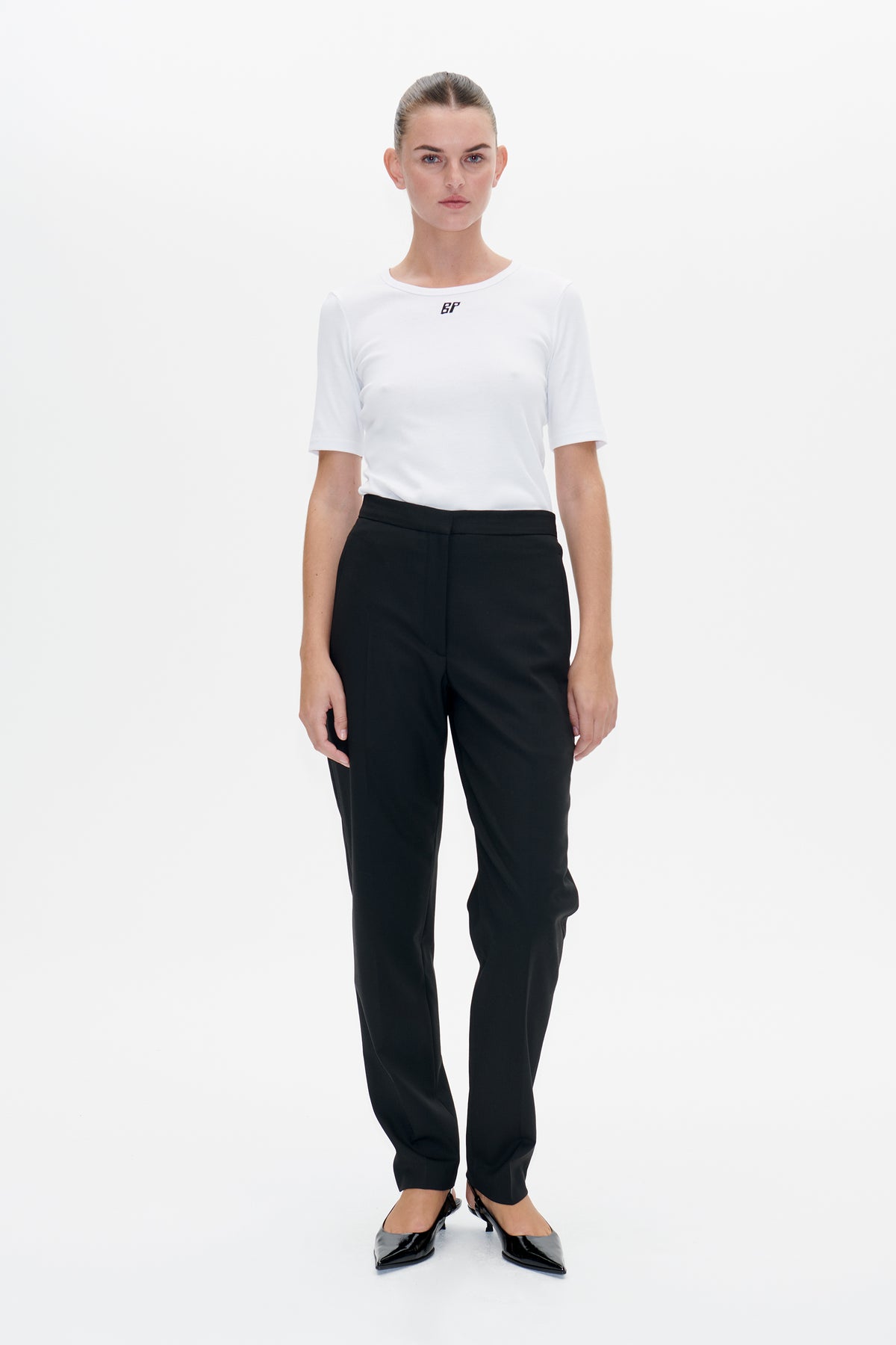 Front view of a model wearing a white short-sleeve T-shirt with a small black chest logo, black trousers and pointed shoes.
