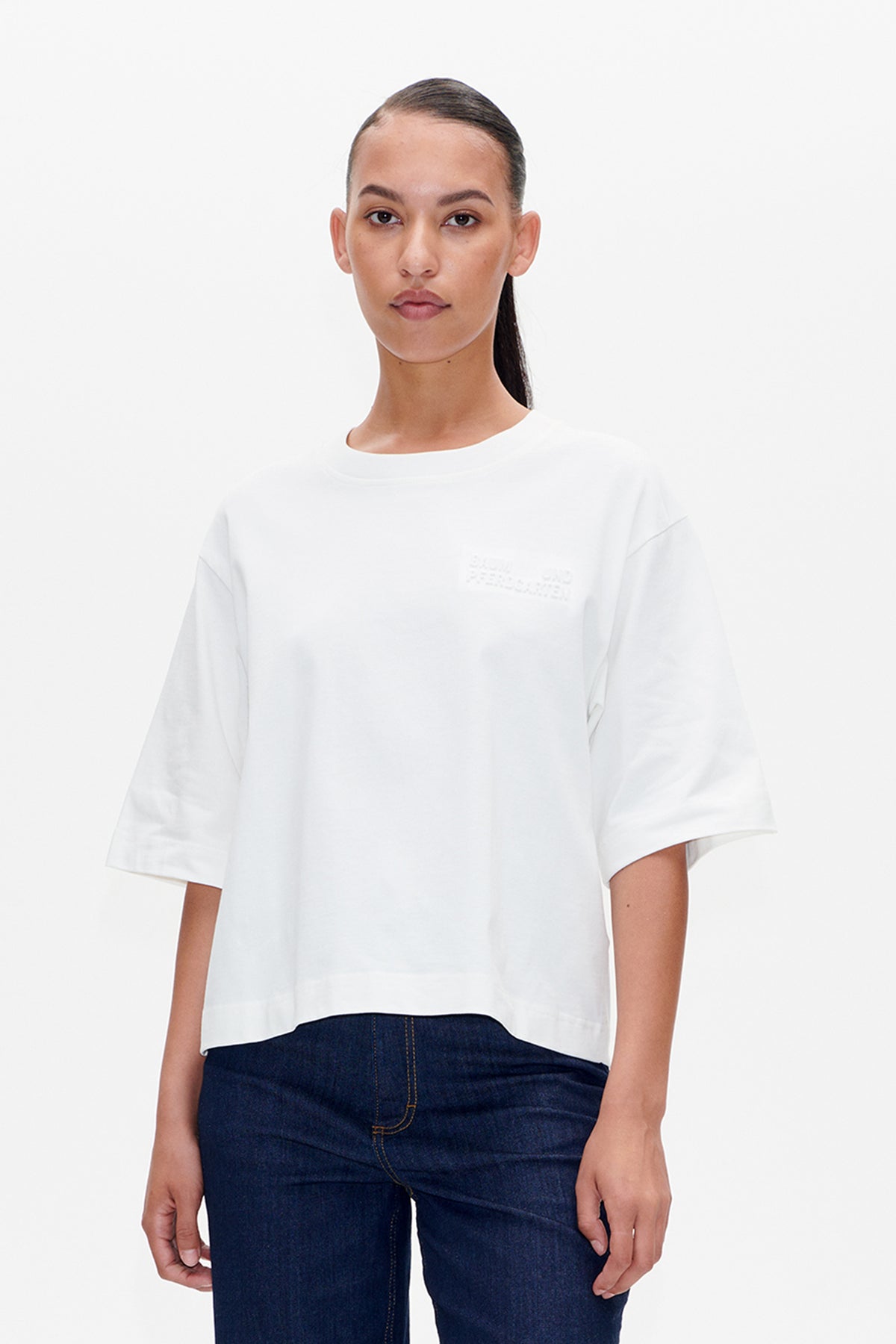 Front view of a white short-sleeved T-shirt with subtle tonal chest lettering, worn with dark indigo jeans.