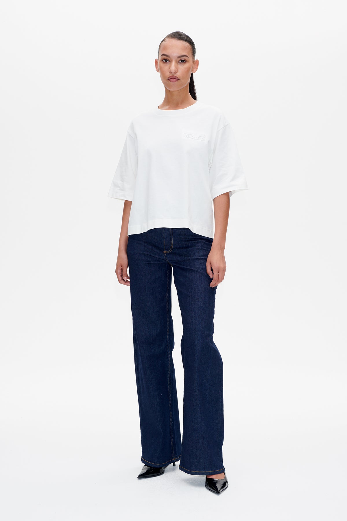 Front view of a model wearing an oversized white short-sleeve T-shirt with a subtle tonal chest detail and dark wide-leg jeans.