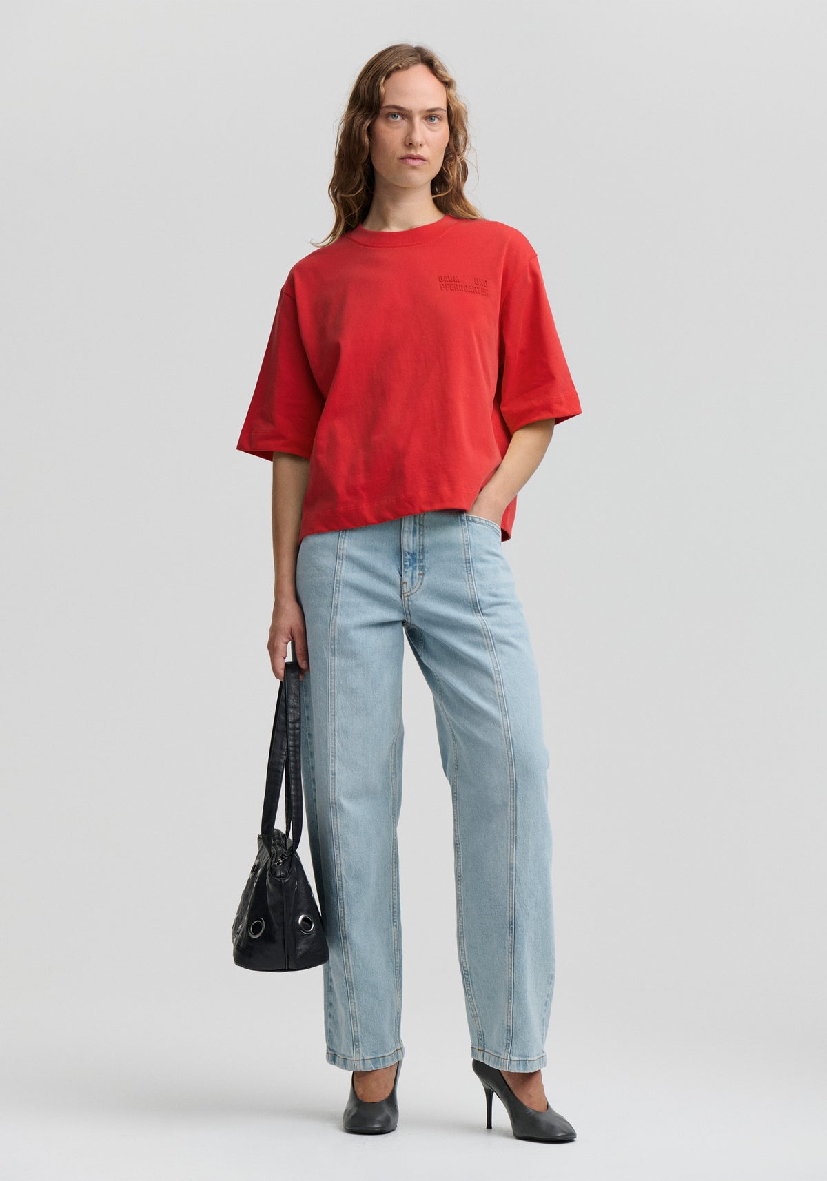 A woman modeling an oversized red short-sleeve t-shirt with a chest pocket, paired with light-wash straight-leg jeans, black heels, and holding a black handbag against a light gray studio background.
