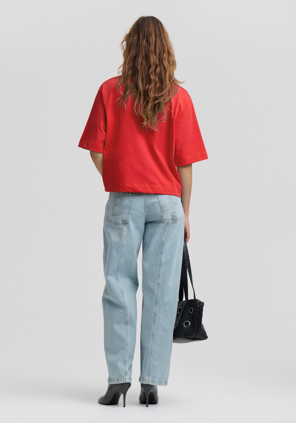 Eine Frau ist von hinten zu sehen. Sie trägt ein knallrotes oversized T-Shirt mit kurzen Ärmeln, hellblau gewaschene Jeans mit weitem Bein und schwarze High Heels. Sie hält eine schwarze Handtasche vor einem hellgrauen Studiohintergrund.