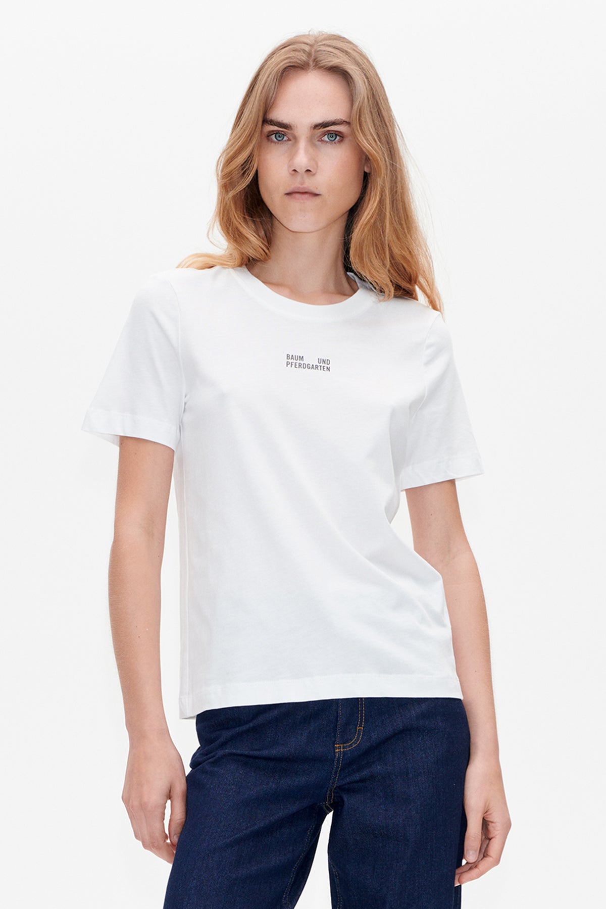Front view of a white short-sleeved T-shirt with a small Baum und Pferdgarten logo, styled with dark indigo jeans.