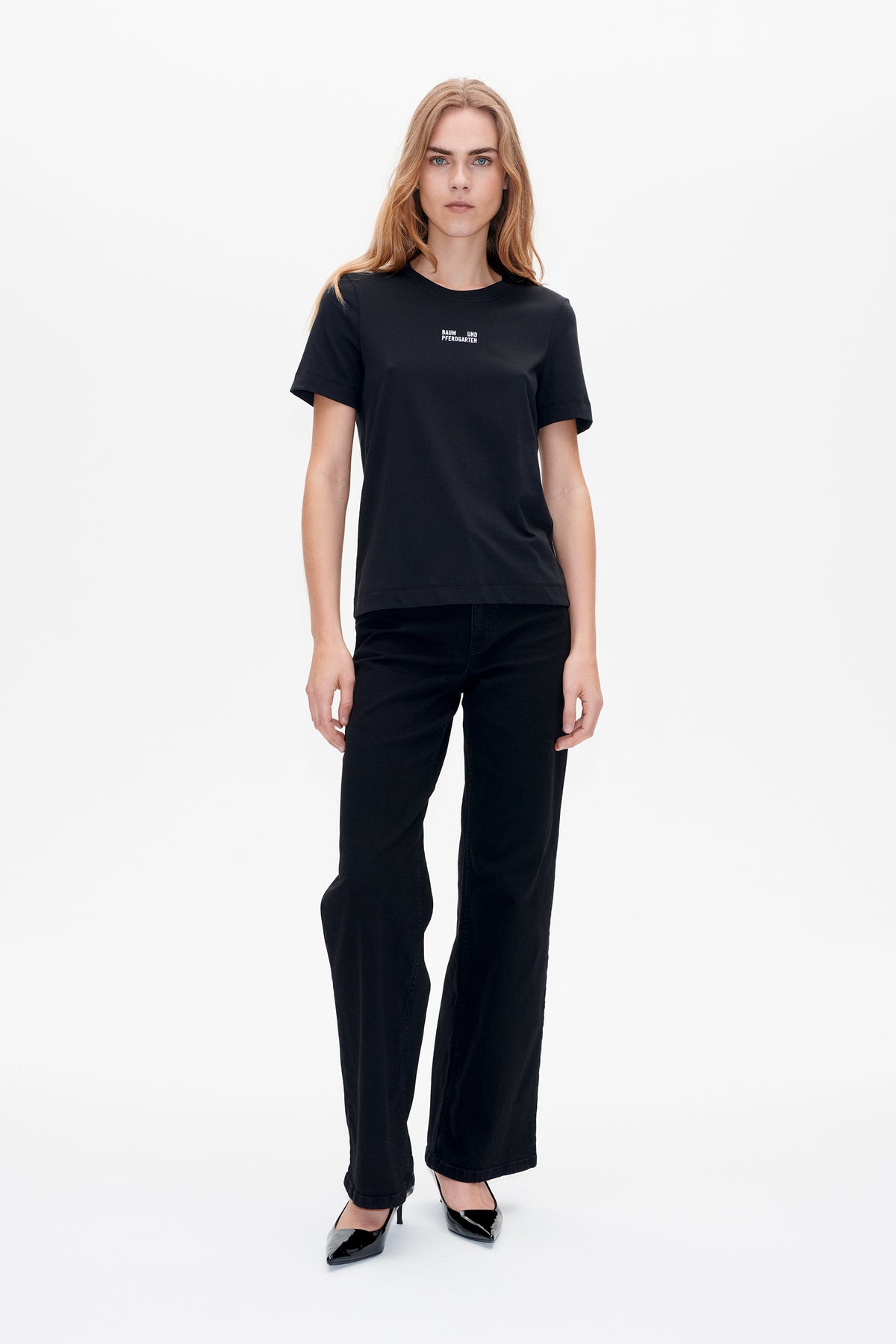Front view of a model wearing a black short-sleeve crew-neck T-shirt with a small white Baum und Pferdgarten chest logo and black wide-leg jeans.