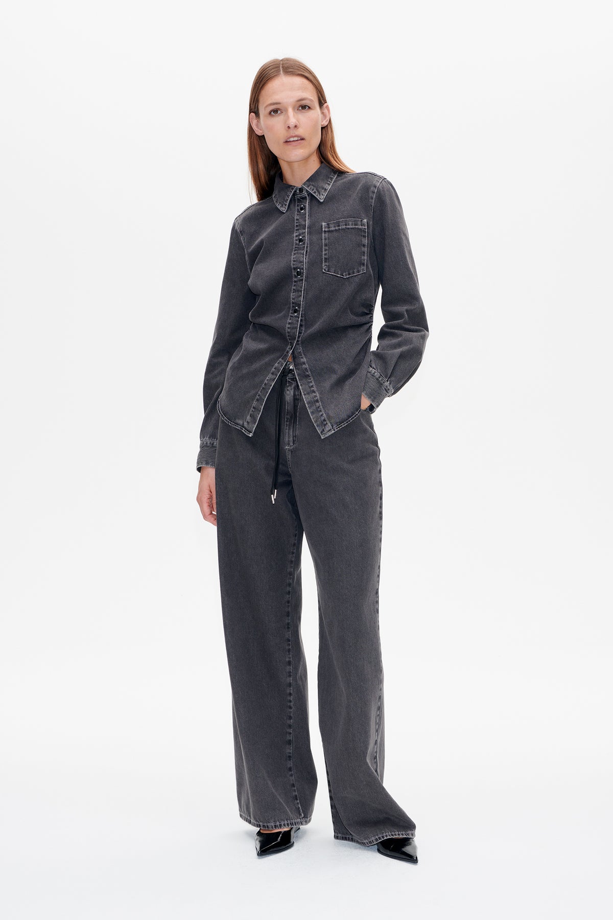 Vooraanzicht van een model dat een antracietgewassen denimoverhemd met knoopsluiting en borstzak draagt, gecombineerd met bijpassende wijde jeans.