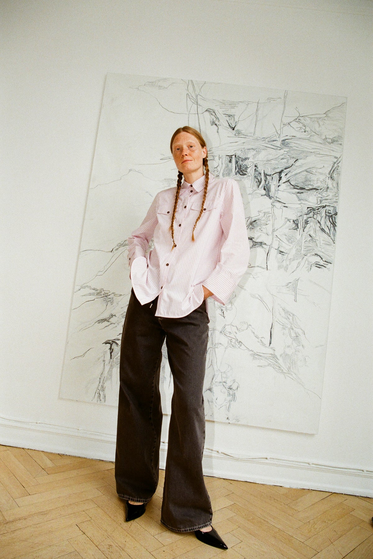 Model draagt een lichtroze, fijngestreepte blouse met borstzakken en een donkere, wijdvallende broek, staand voor een grote schets.