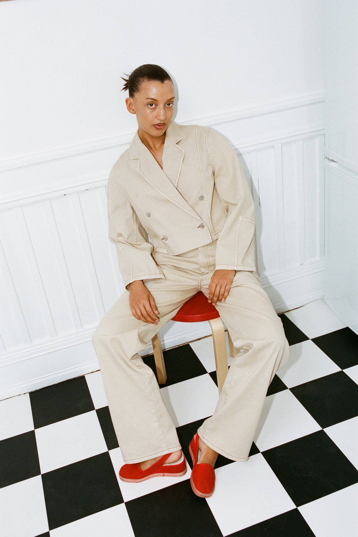 Mannequin assis contre un mur blanc à panneaux, portant une veste crème aux surpiqûres contrastantes et un pantalon large, avec des chaussures rouge vif.