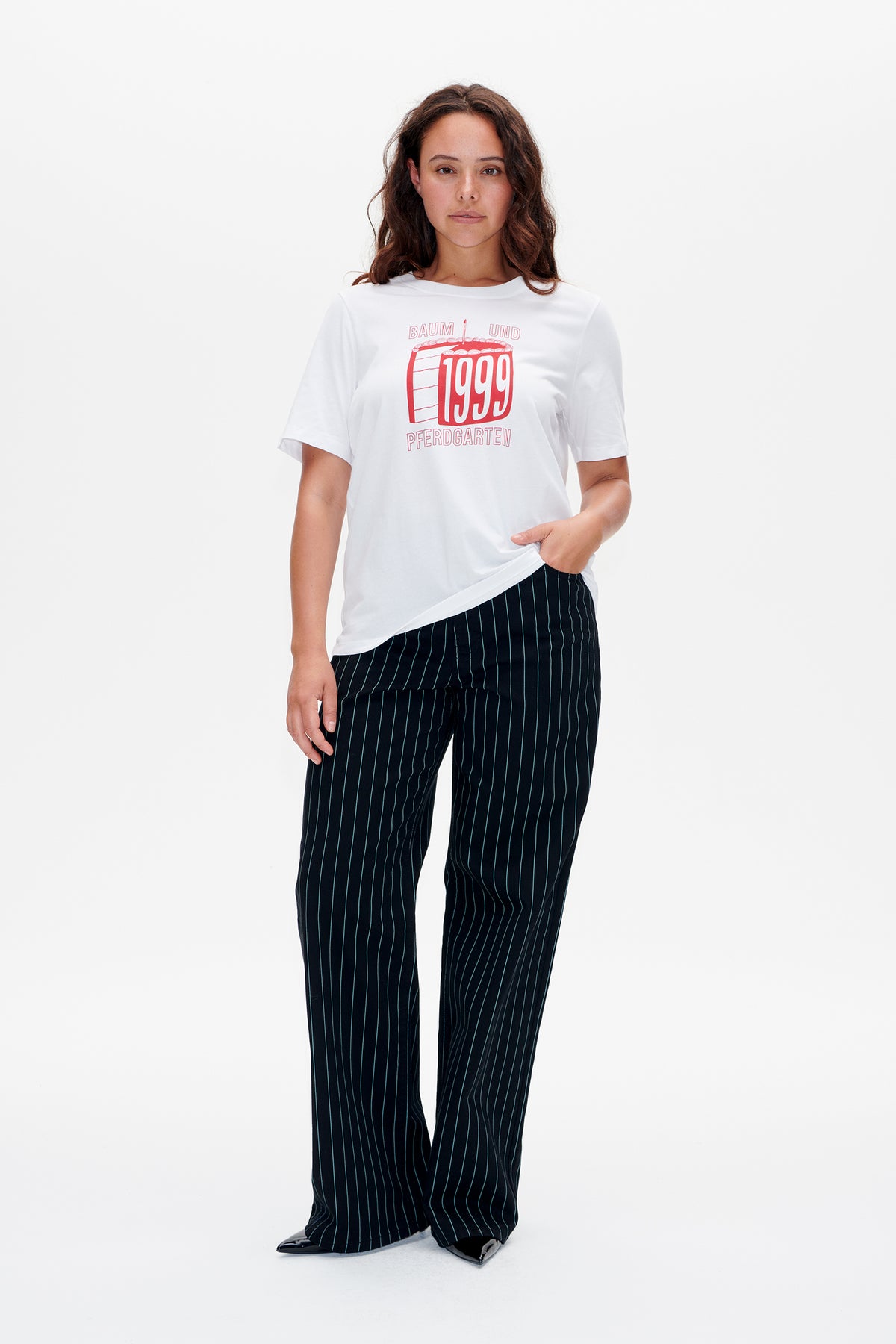 Model draagt een wit T-shirt met een rode '1999' taartprint en een zwarte broek met dunne verticale strepen.