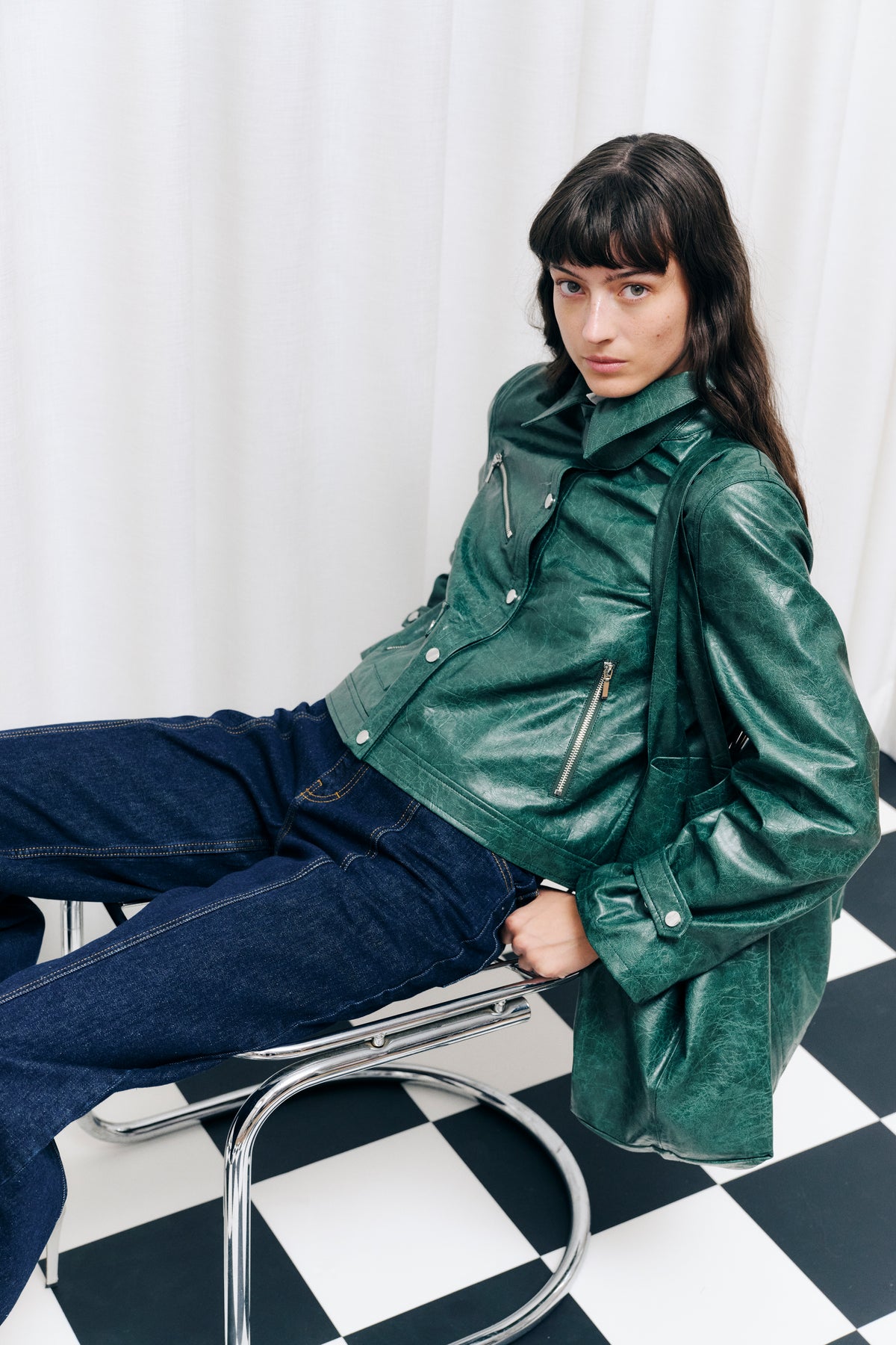 Campaign portrait of a seated model wearing a crinkled dark green jacket with a wide collar and silver zips, paired with dark blue jeans.