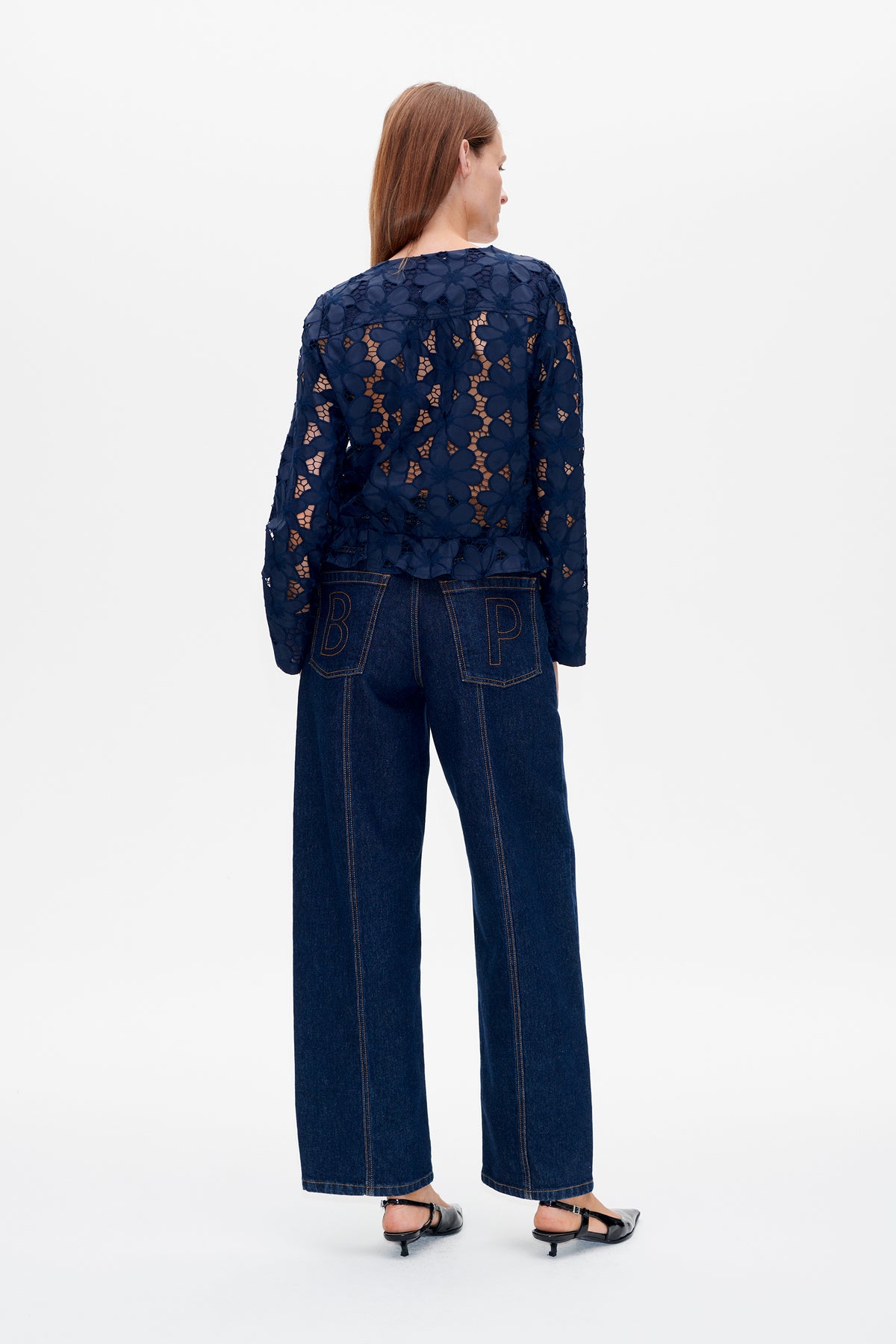Rückansicht eines Models in einer marineblauen Spitzenjacke mit Blumenmuster und Schößchen sowie dunklen, weitgeschnittenen Jeans.