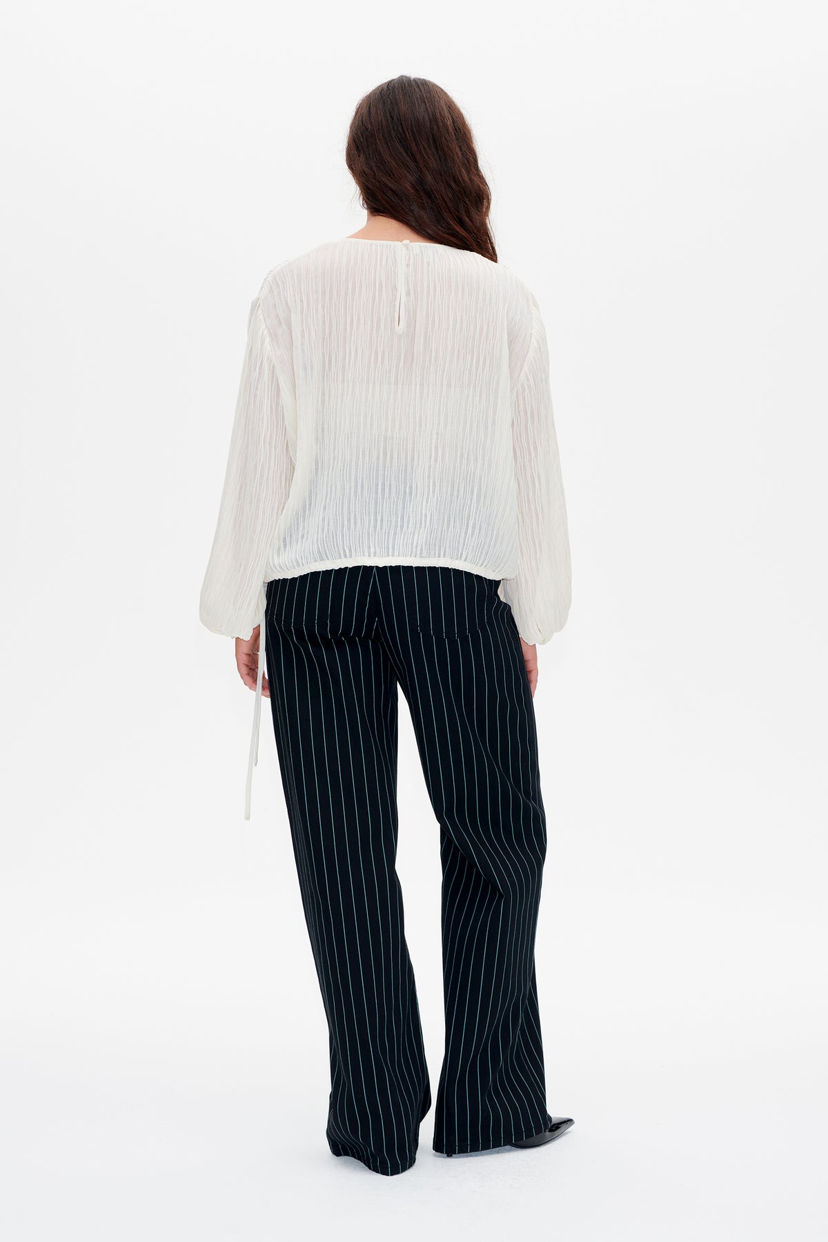 Back view of a model in an ivory long-sleeved blouse with crinkled vertical texture and dark wide-leg trousers with thin pinstripes.