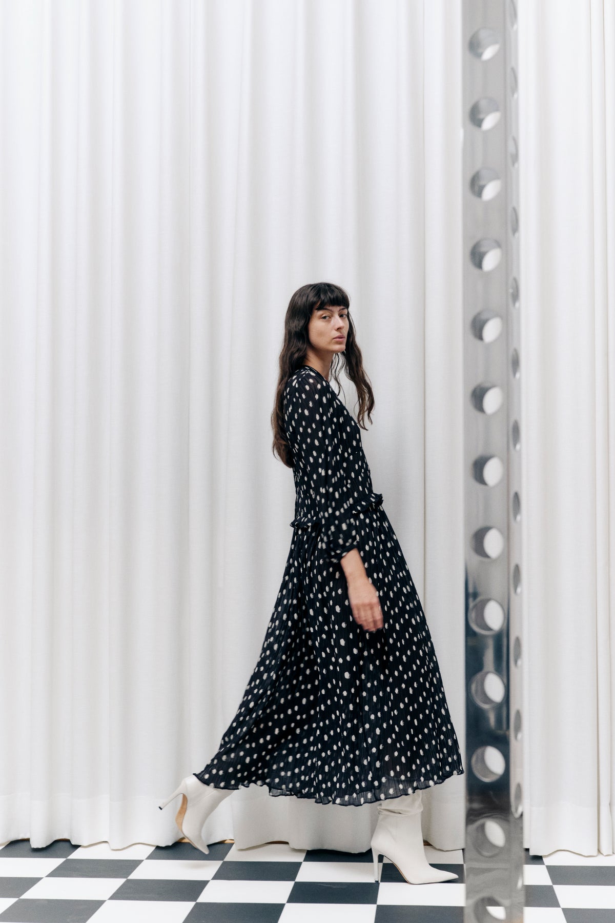 Model poses in a flowing black dress with irregular cream spots and ruffled tiers against white curtains and a checkerboard floor.