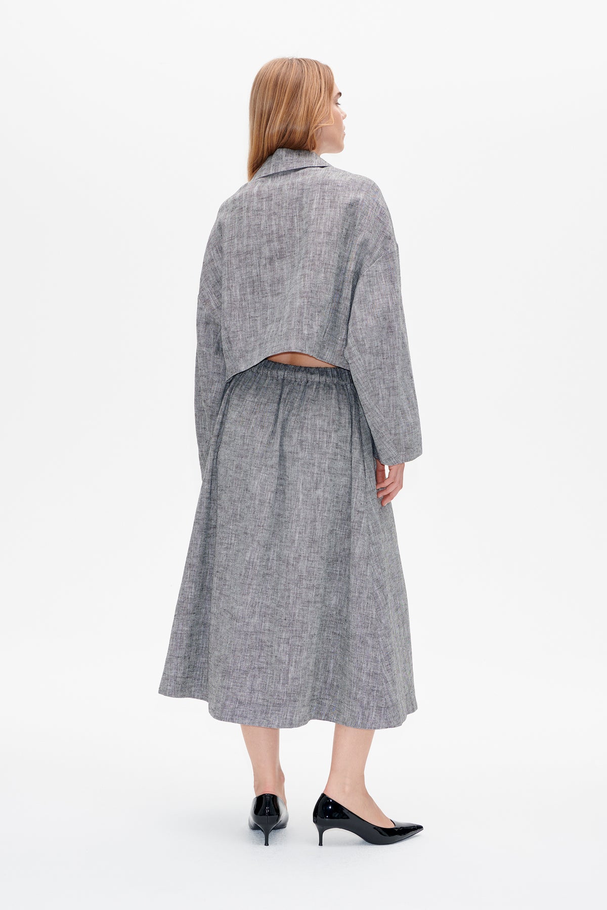 Back view of a model wearing a cropped grey herringbone jacket and matching midi skirt with an elasticated waist.