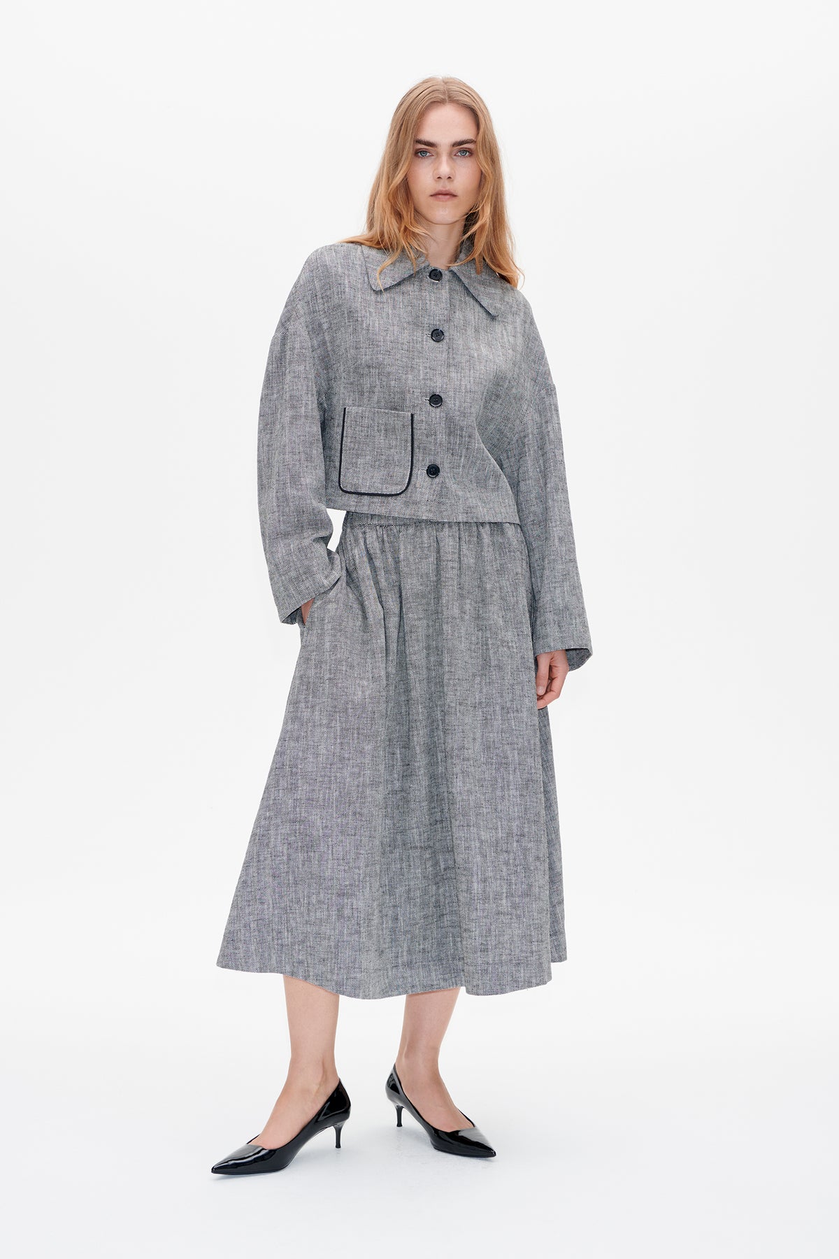 Model wearing a grey herringbone button-front jacket with a large piped pocket and a matching pleated midi skirt.