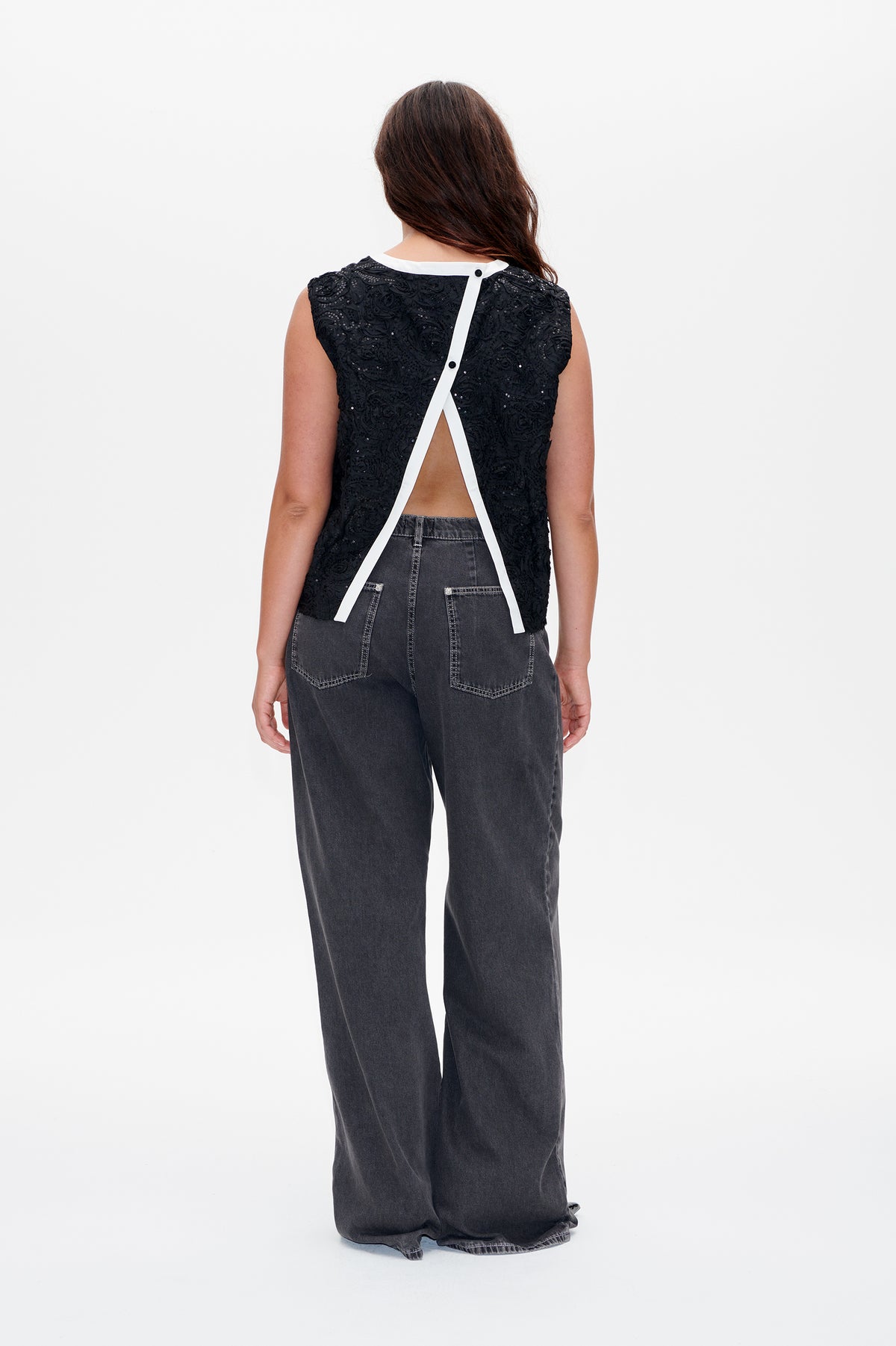 Rückansicht eines schwarzen, ärmellosen Tops mit texturierten Blumenapplikationen, weißem, überkreuztem Rückendetail und offenem Einsatz, getragen zu anthrazitfarbenen, weit geschnittenen Jeans.