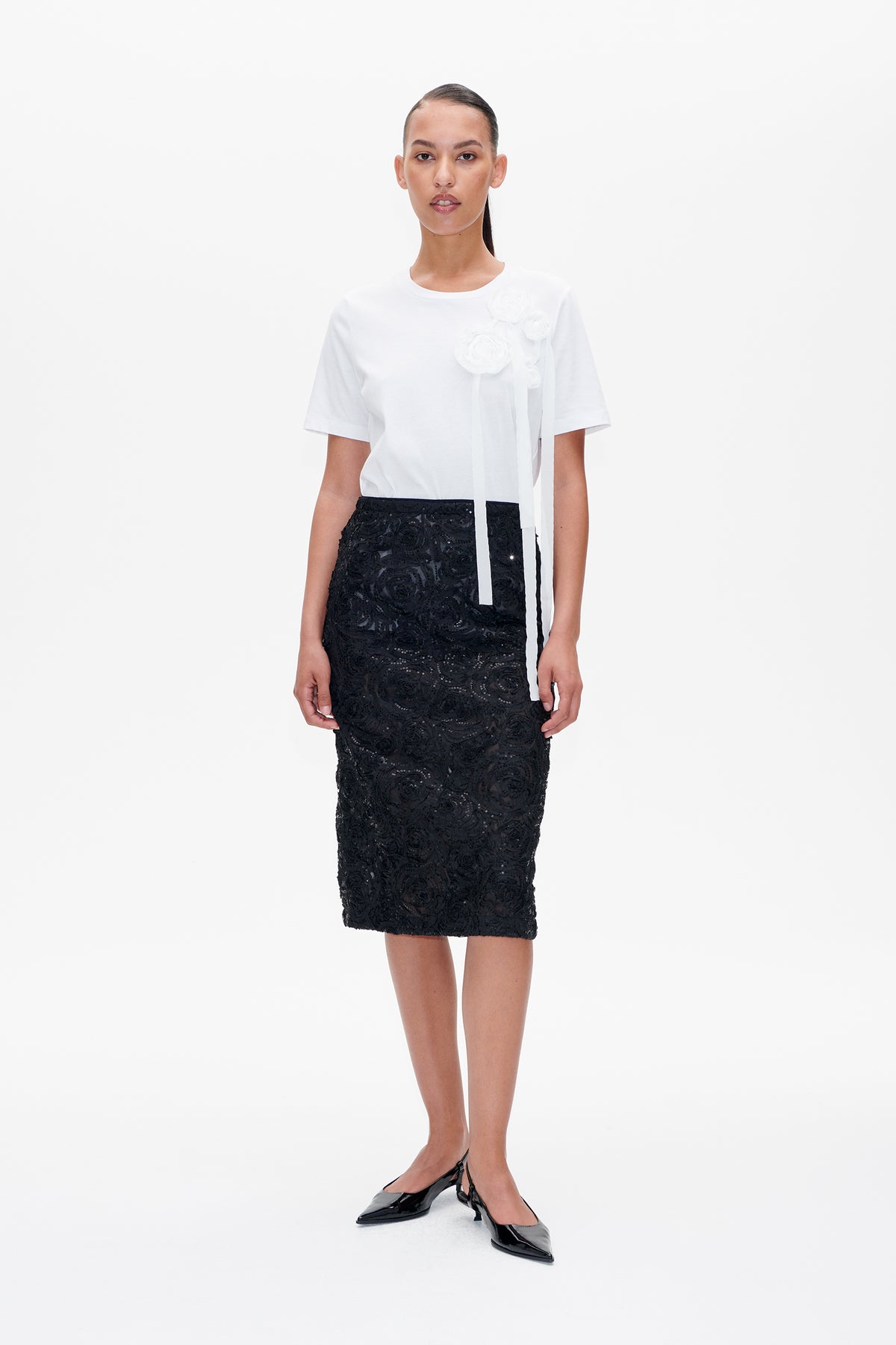 Front view of a model wearing a white short-sleeved T-shirt with fabric flowers and long ribbon ties, paired with a black sequined lace midi skirt.