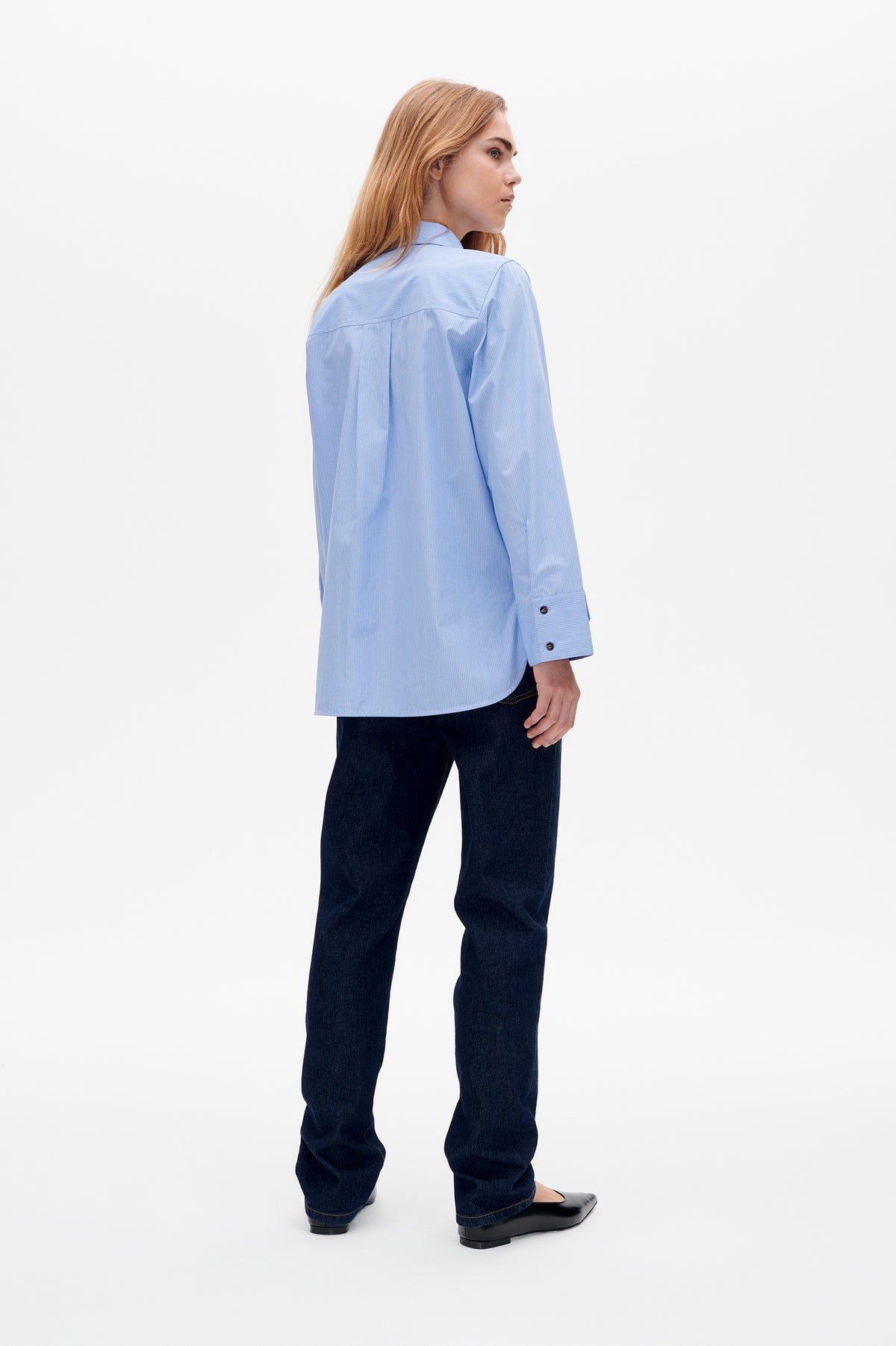 Model wearing a light blue fine-striped shirt, shown from a three-quarter back view with a back yoke and pleat, paired with dark jeans.