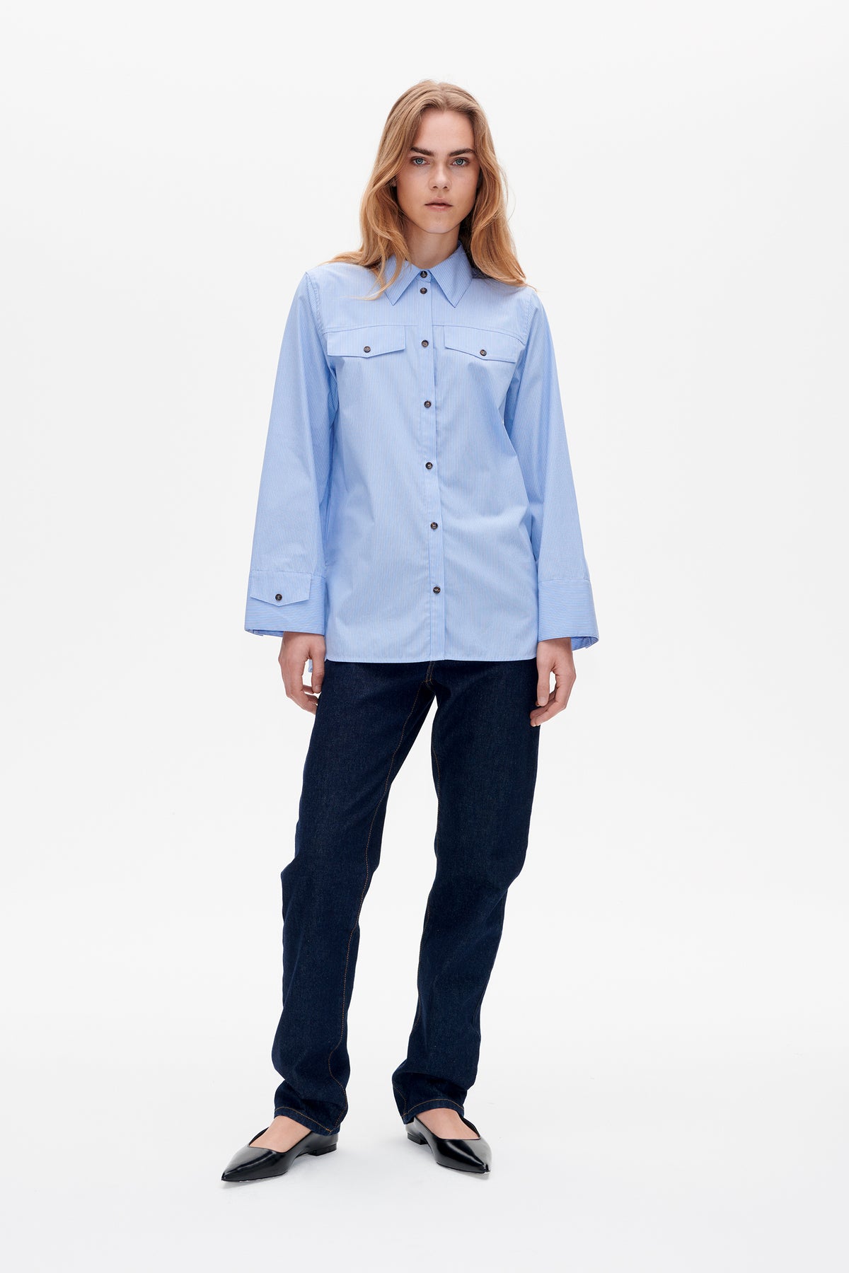 Front view of a model wearing a light blue pinstriped button-up shirt with chest pockets and wide cuffs, dark jeans and black flats.