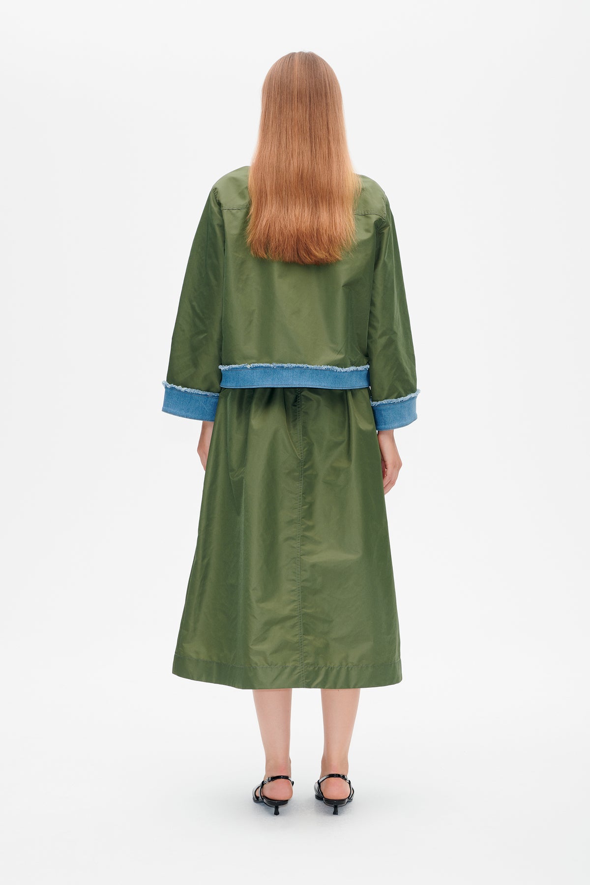 Back view of a model wearing a knee-length olive green coat with wide blue denim cuffs and matching denim waist trim.