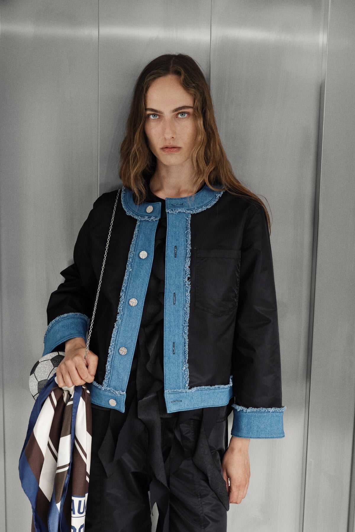 Model in a black jacket with blue denim panels and frayed trim, holding a blue, brown and cream geometric scarf against a grey wall.