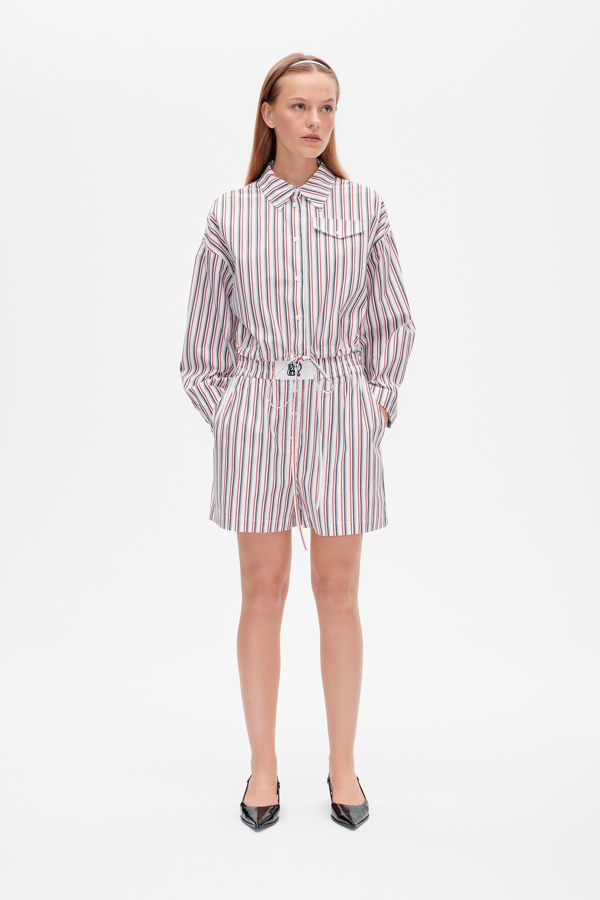 Model trägt weiße, gestreifte Hemdbluse mit Knopfleiste, Brusttasche und Taillenzug, kombiniert mit passender Shorts.