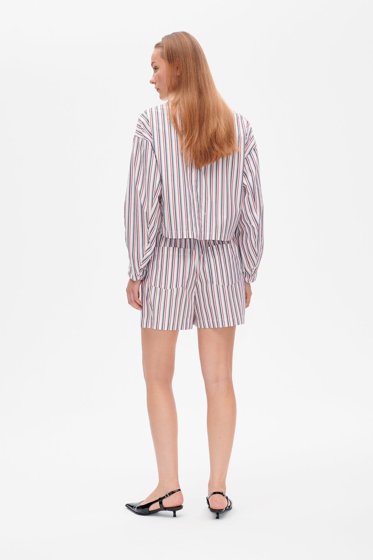 Rückenansicht eines Models, das ein weißes Langarmtop und Shorts mit roten, marineblauen und grauen vertikalen Streifen trägt.