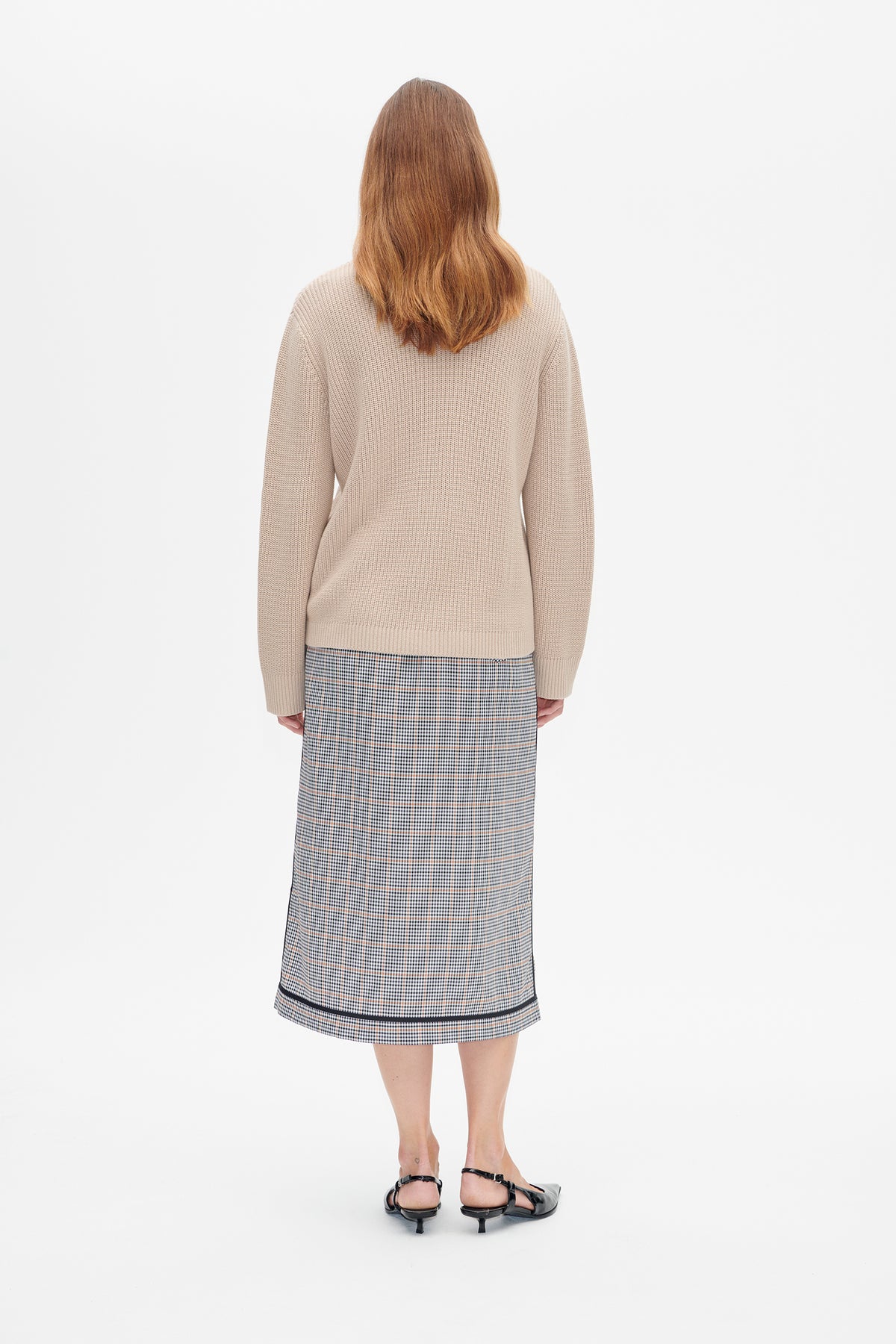 Back view of a model wearing a pale beige textured knit sweater and a grey checked midi skirt with thin orange lines.