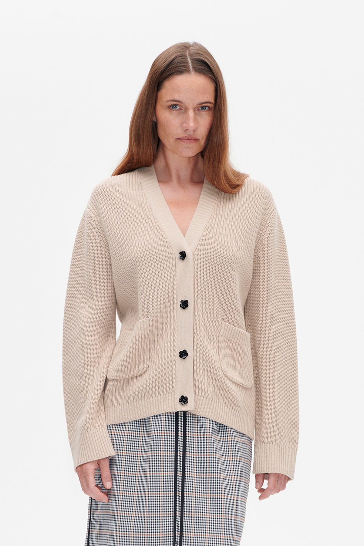 Front detail of a light beige knit cardigan with black flower-shaped buttons and patch pockets, worn over a black-and-white checked garment.