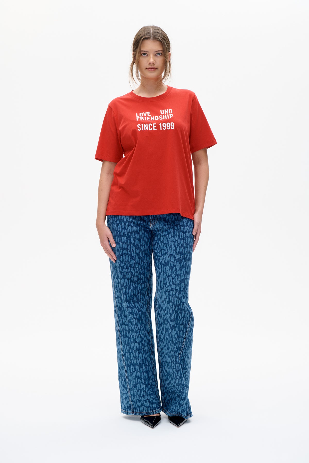 Model wearing a red short-sleeved T-shirt printed “LOVE UND FRIENDSHIP SINCE 1999” with blue patterned wide-leg jeans.