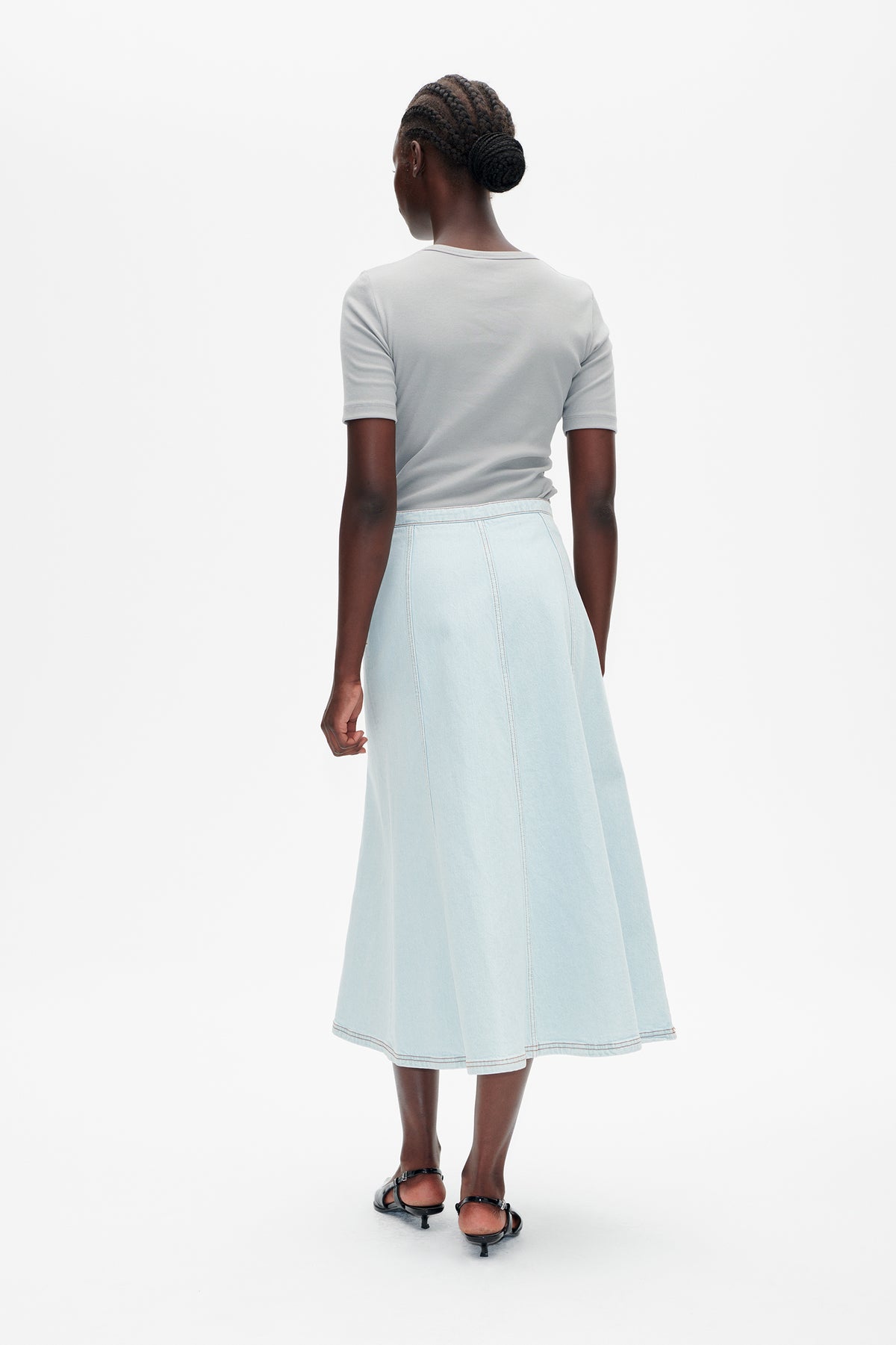 Achteraanzicht van een model in een lichtblauwe denim midi-rok met paneelnaden en een lichtgrijze top met korte mouwen.