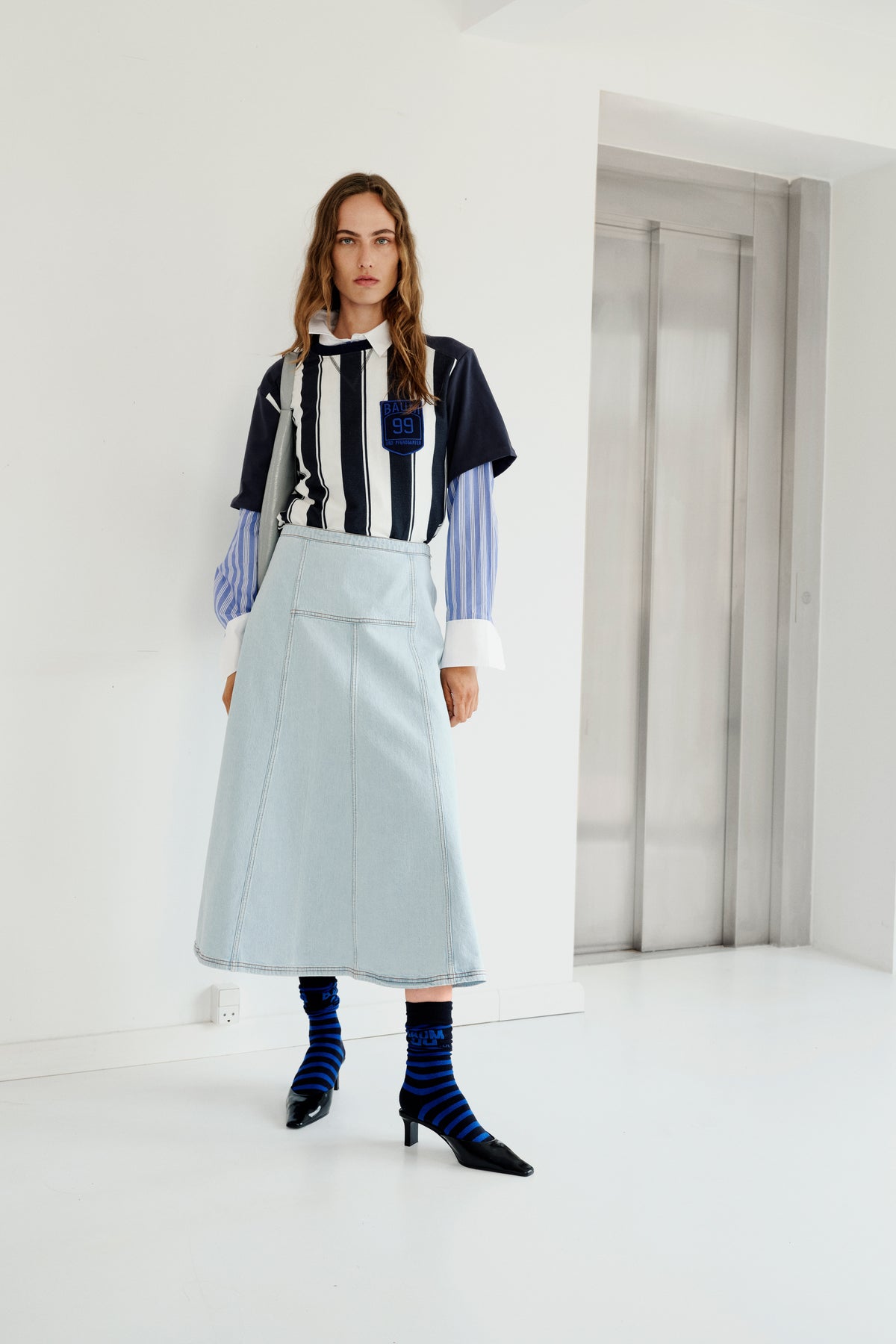 Model in een lichte, minimalistische ruimte draagt een marineblauw-wit gestreepte top met korte mouwen over een blauw gestreepte blouse, een lichte denimrok en gestreepte sokken.