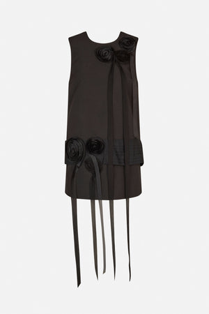 Front view of a black sleeveless dress with sculptural rosette details, pleated hem panels and long hanging ribbons.