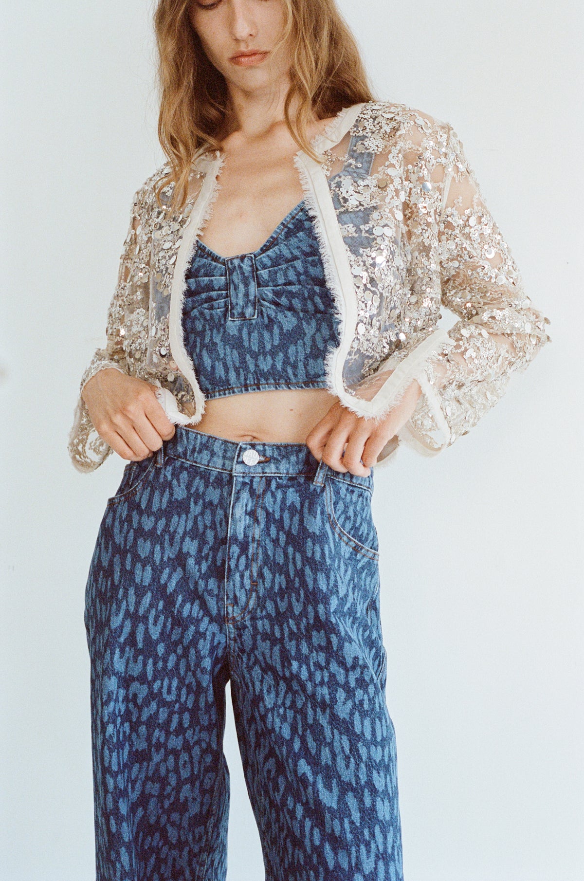 Campaign portrait of a model in a sheer beige sequin jacket over a blue denim top and matching patterned jeans.