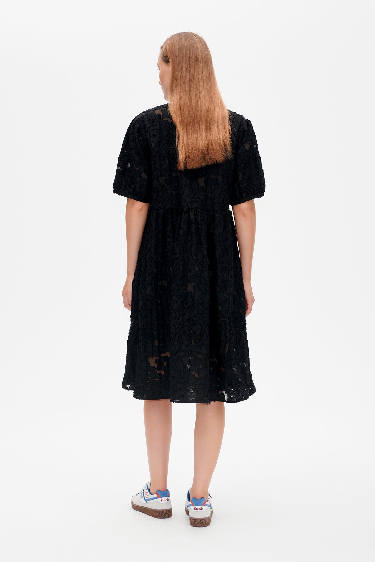 Back view of a black knee-length dress with short sleeves, a gathered waist and textured floral lace pattern.