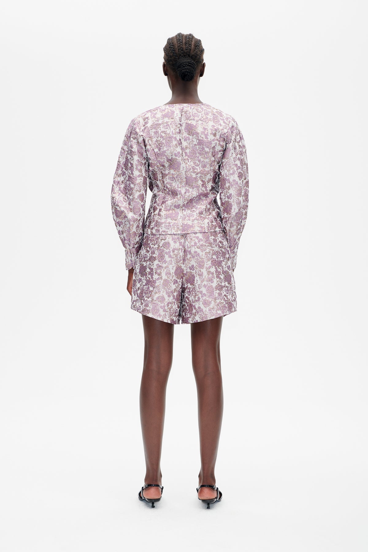 Back view of a model wearing a lilac and cream floral jacket with matching tailored shorts and black strappy heels.