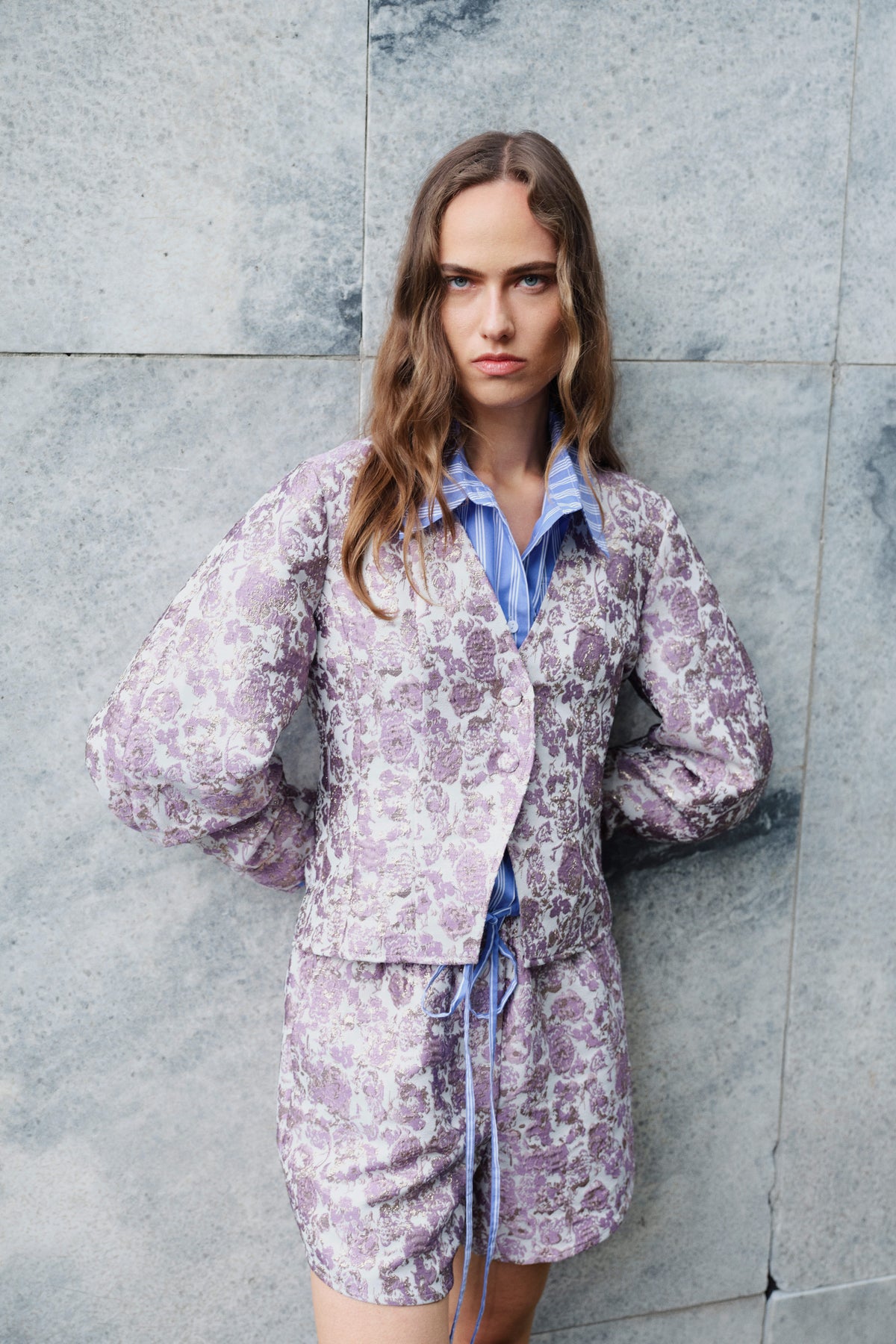 Campaign portrait of a model in a lilac-and-cream patterned blazer and matching skirt over a blue striped collared shirt, against a stone wall.
